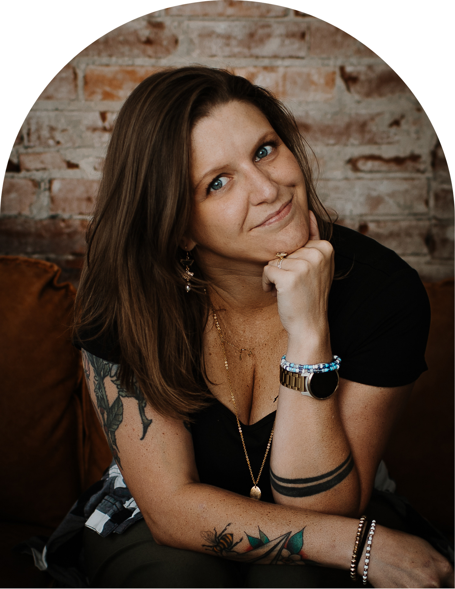 Smiling woman with long brown hair and tattoos rests her chin on her hand while seated on a brown leather couch before a brick wall.