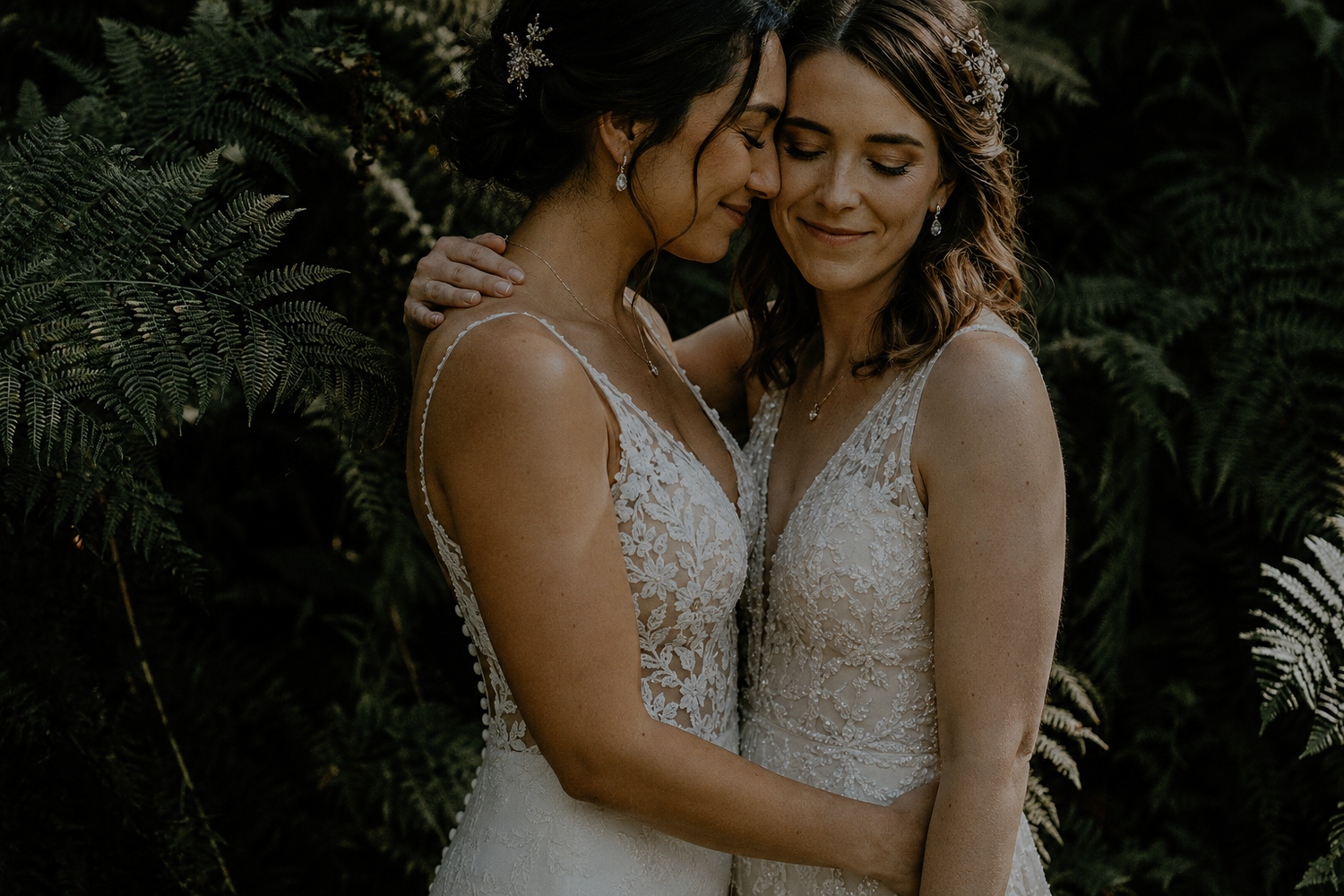 Two brides in lace wedding gowns embrace tenderly among lush green ferns in a romantic outdoor ceremony.