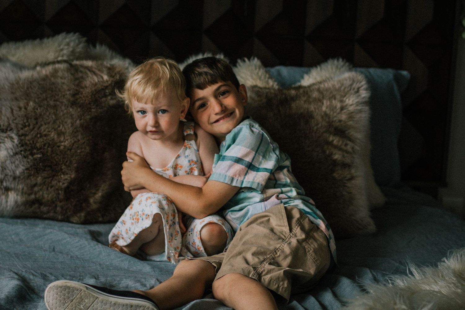 Older brother hugging younger blonde toddler sister on cozy fur-covered bed.