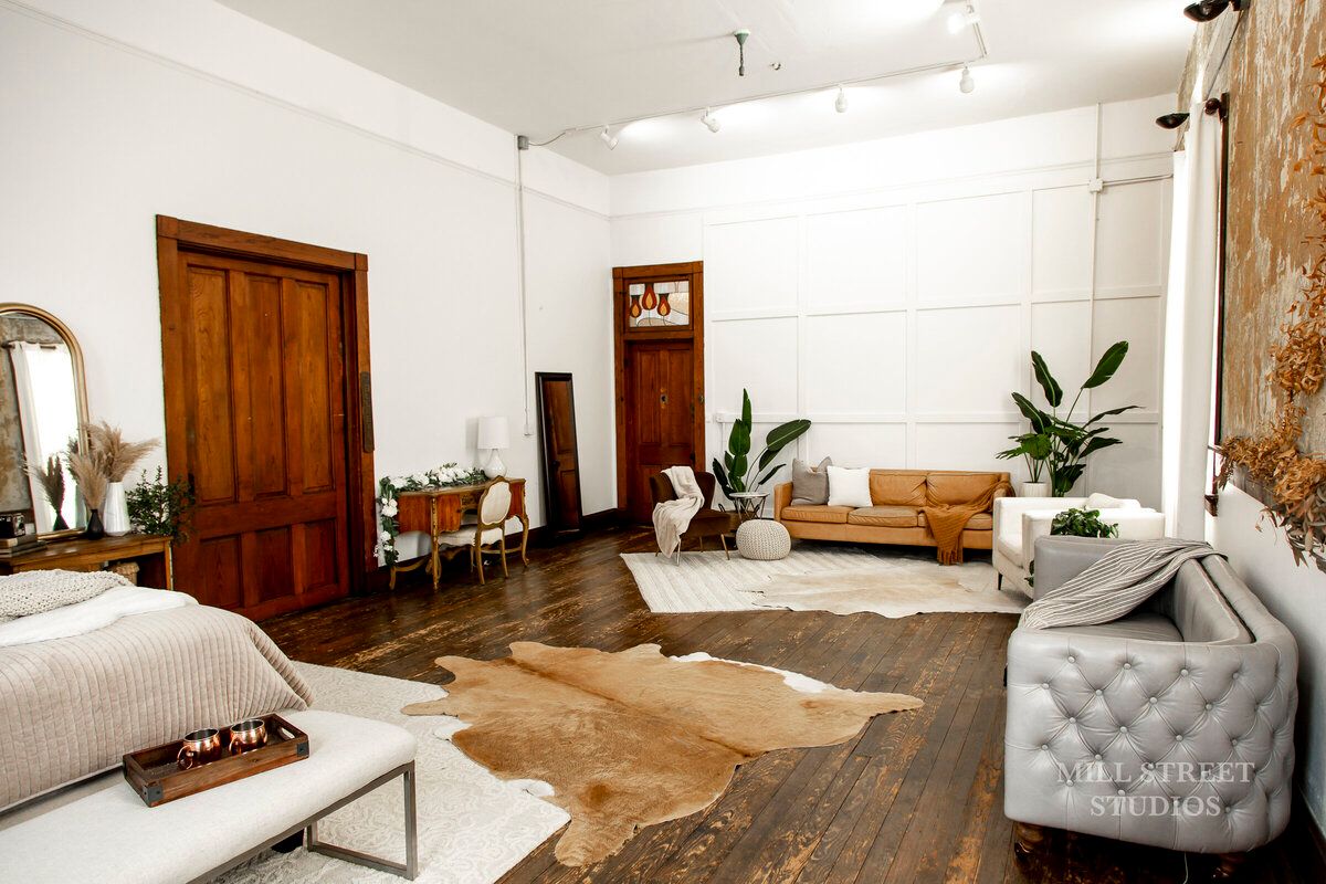 Open boho studio space with leather sofa, tufted chair, cowhide rug, plants, and warm wood floors.