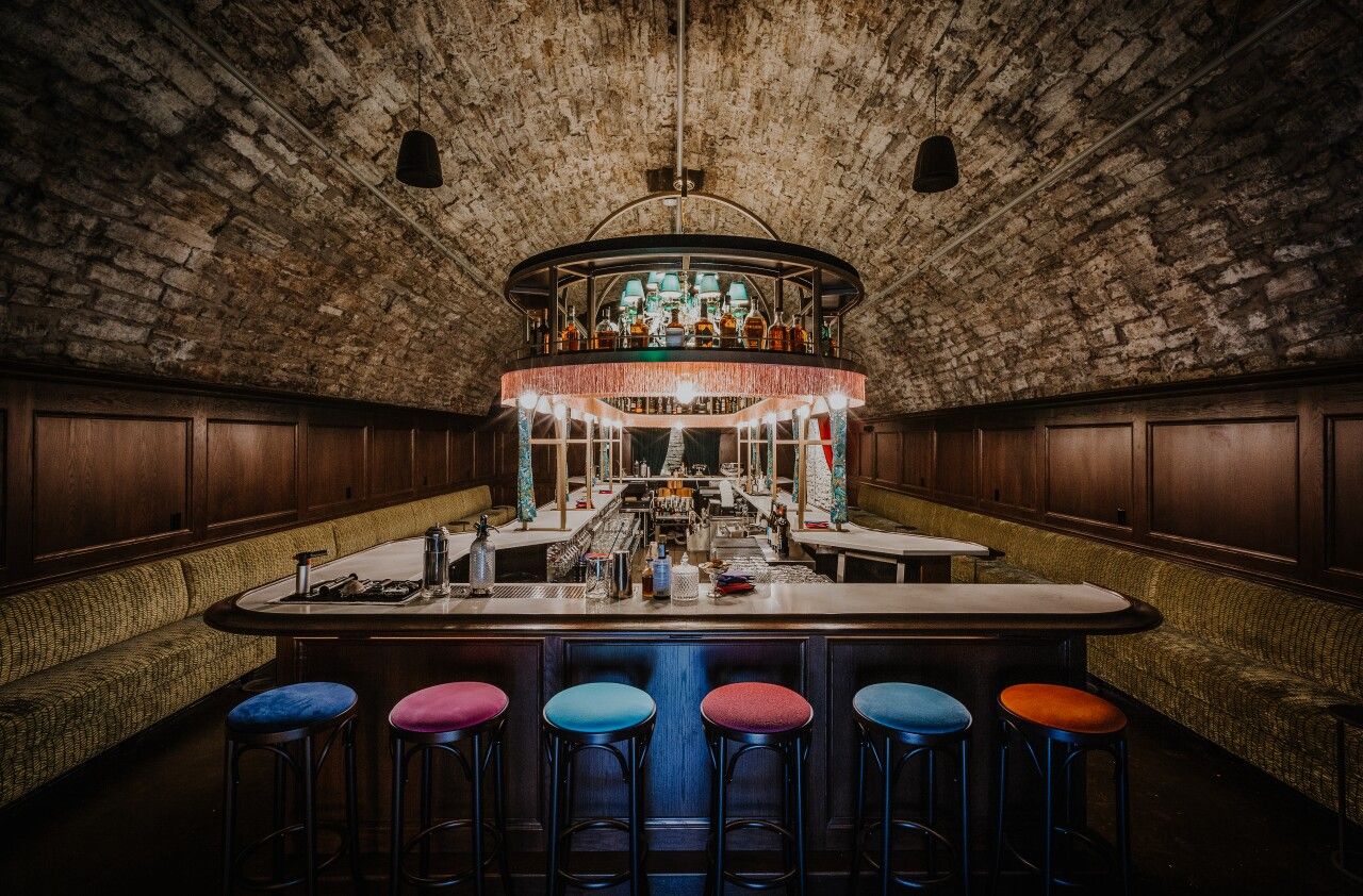 Elegant underground bar with brick vaulted ceiling, colorful stools, dark wood paneling, and glowing chandelier centerpiece.