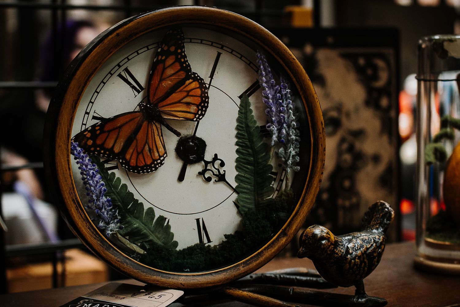 Decorative clock featuring a monarch butterfly and fern design with a small bird figurine on a wooden table.