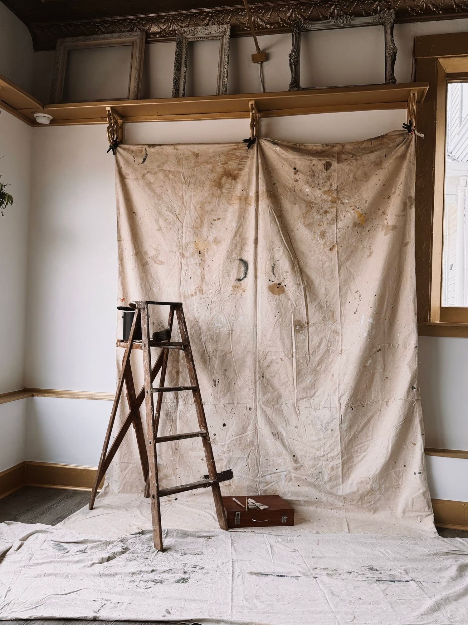 Rustic room renovation with paint-stained drop cloth, wooden ladder, and ornate ceiling detail.