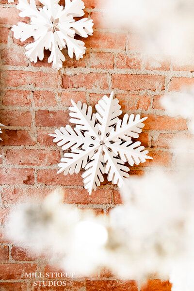 White decorative snowflakes hanging against a rustic red brick wall with soft bokeh snow effect.
