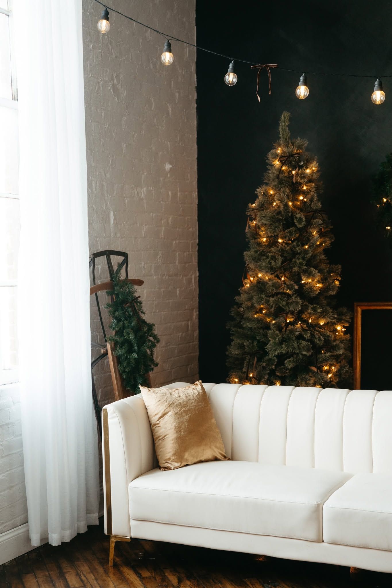 Elegant living room with white tufted sofa, gold pillow, decorated Christmas tree with lights, and string bulb lights on dark wall.