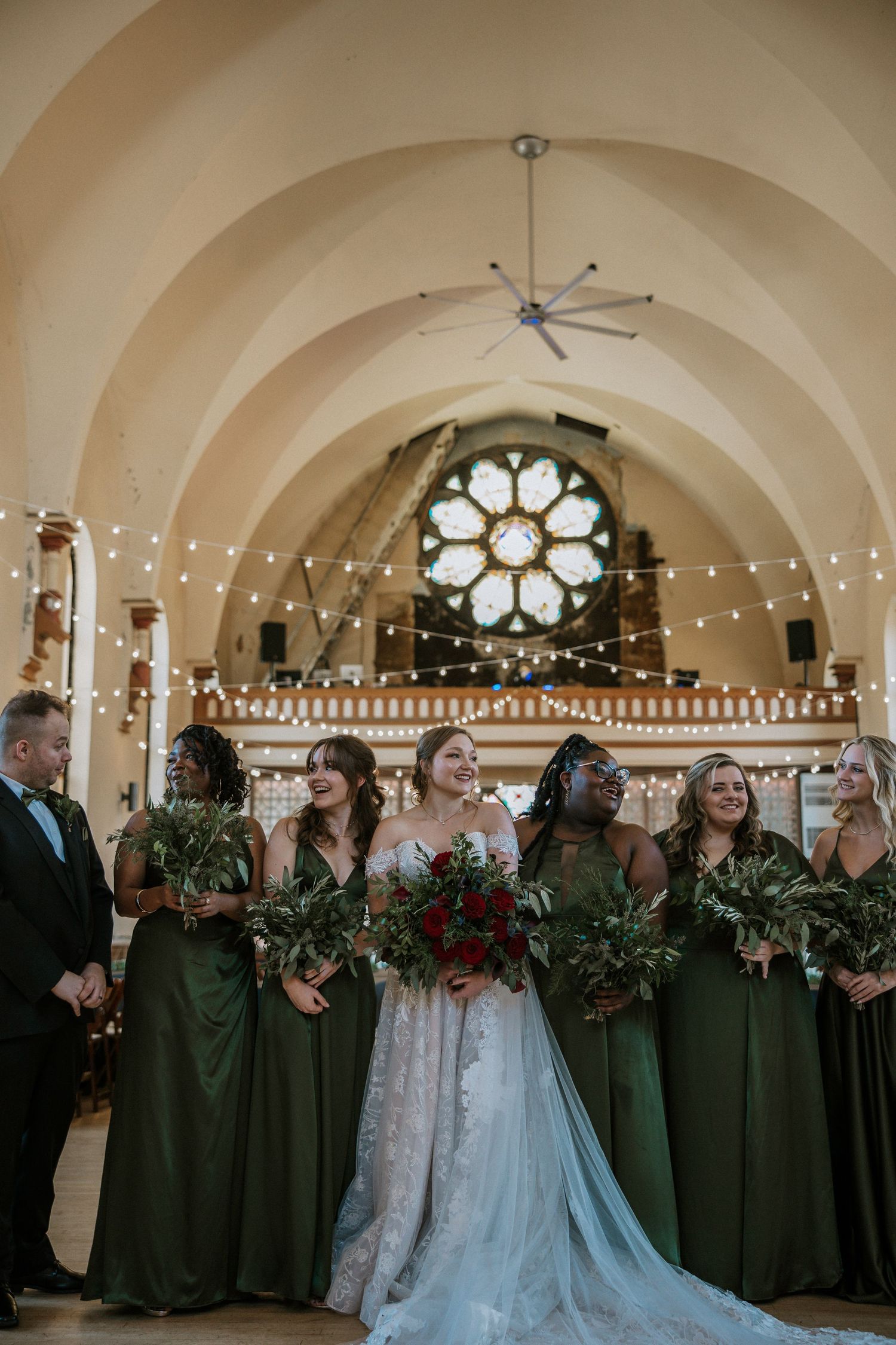 Bride in white lace gown stands with bridesmaids in emerald green dresses inside an elegant church venue.