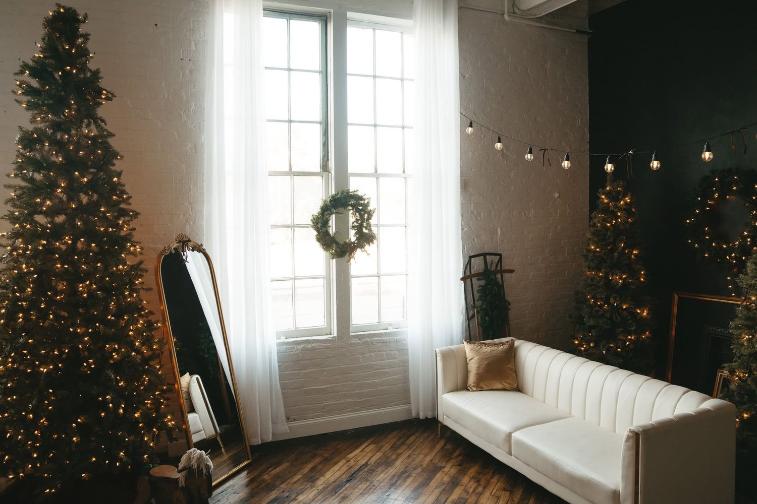 Cozy Christmas living room with two decorated trees, white sofa, wreath on window, and warm string lights.