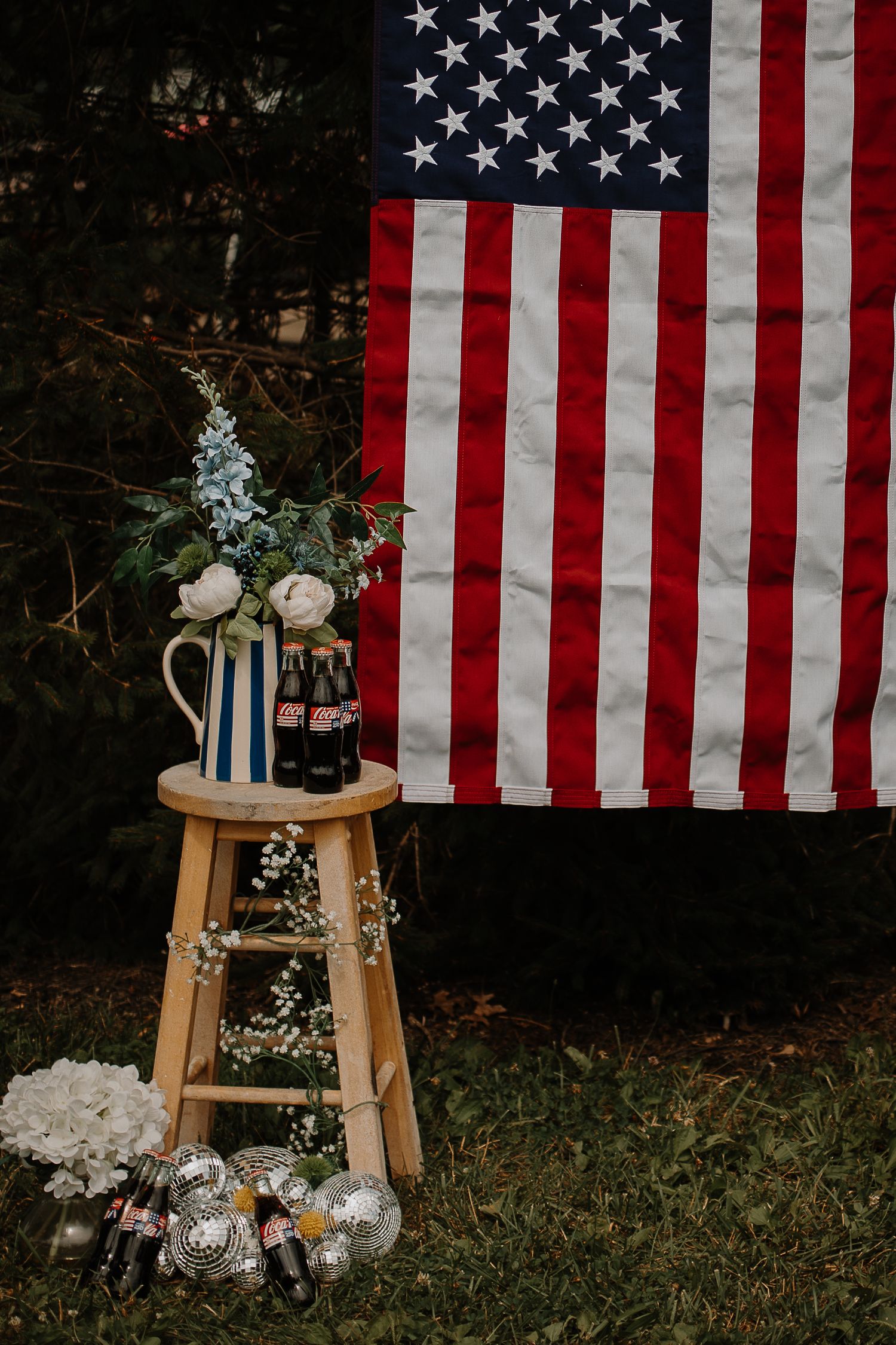 American flag backdrop with floral arrangement, Coca-Cola bottles, and disco balls on a stool.