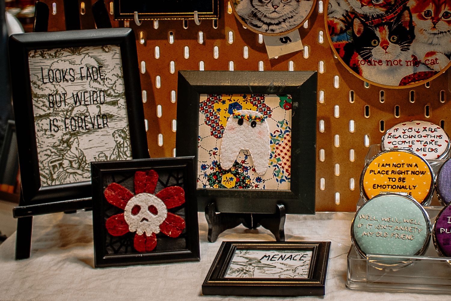 Framed embroidery and mixed media art pieces displayed at an indie craft market.