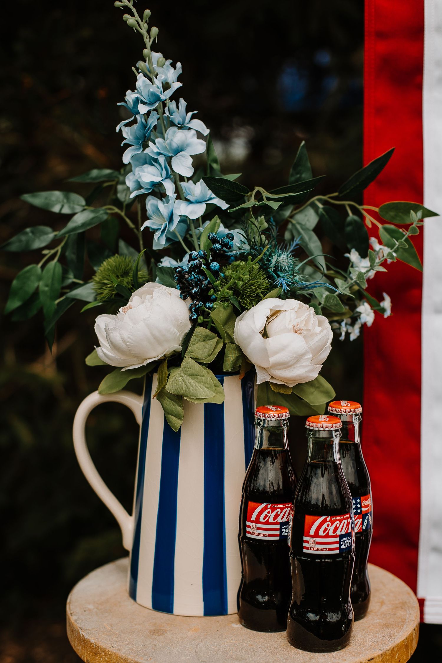 Blue striped pitcher with white peonies and blue flowers beside vintage Coca-Cola bottles on wooden stump.