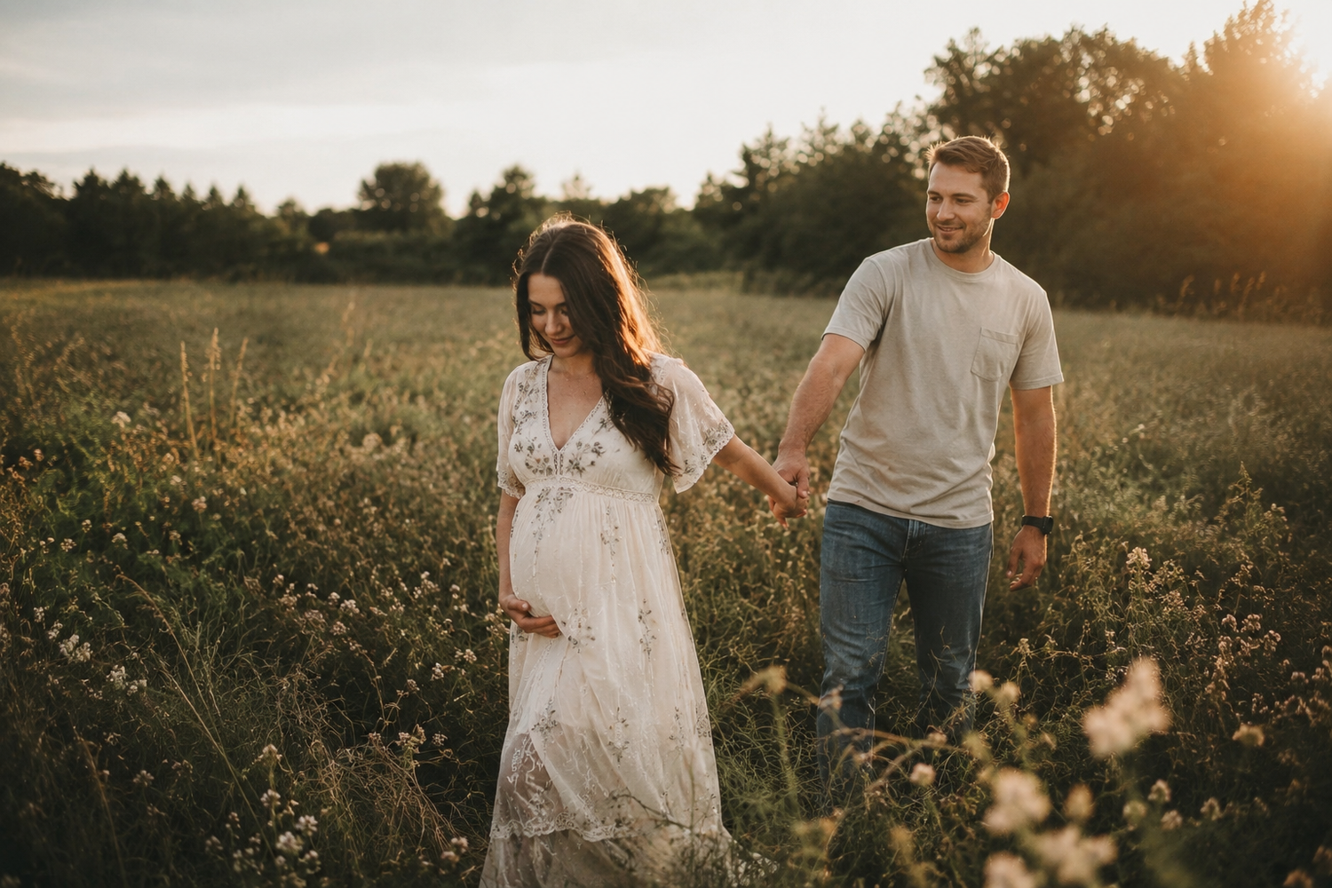 Pregnant woman in white lace dress holds hands with man in field at golden hour sunset.