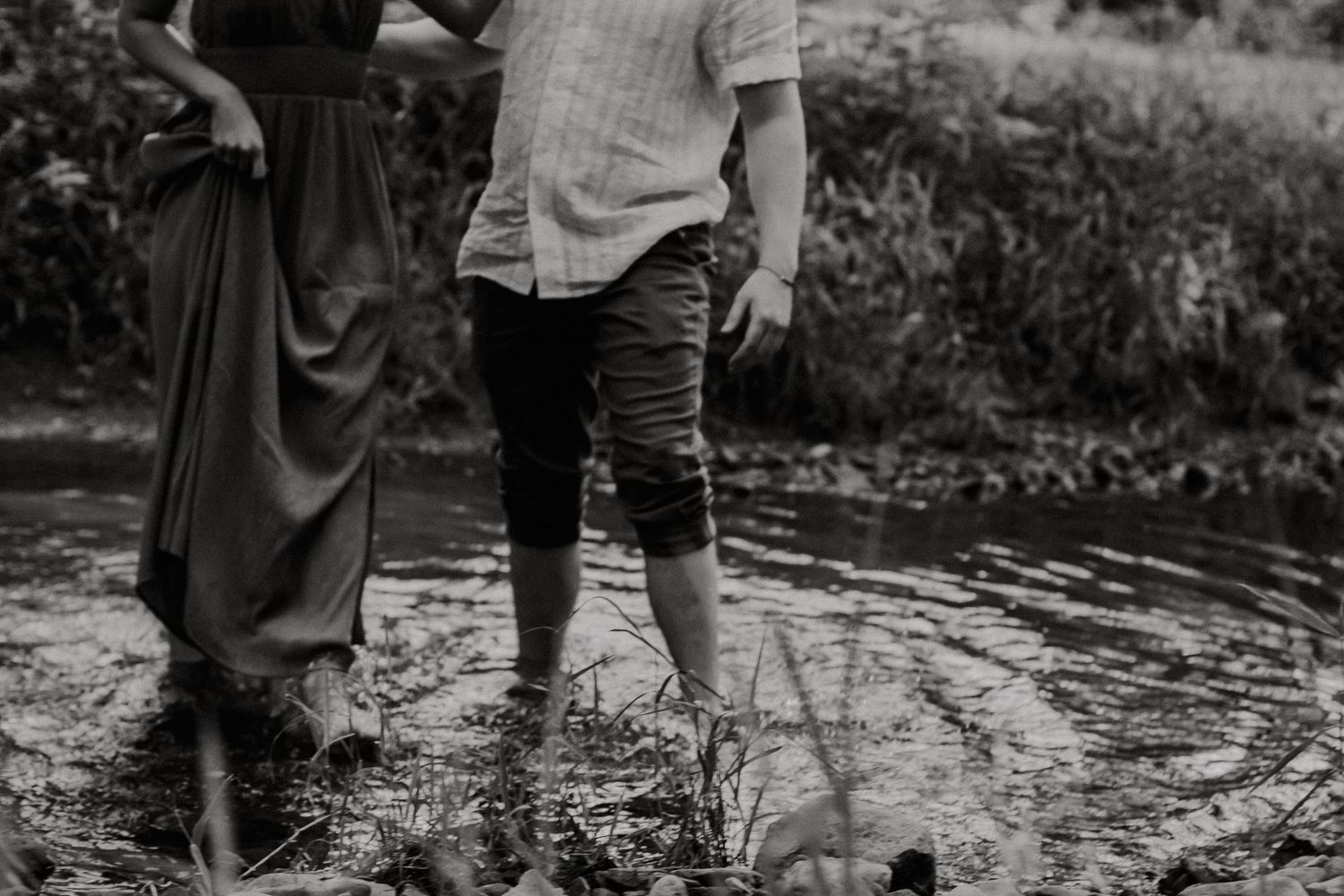 Black and white photo of barefoot couple standing together in a shallow creek during outdoor engagement session.