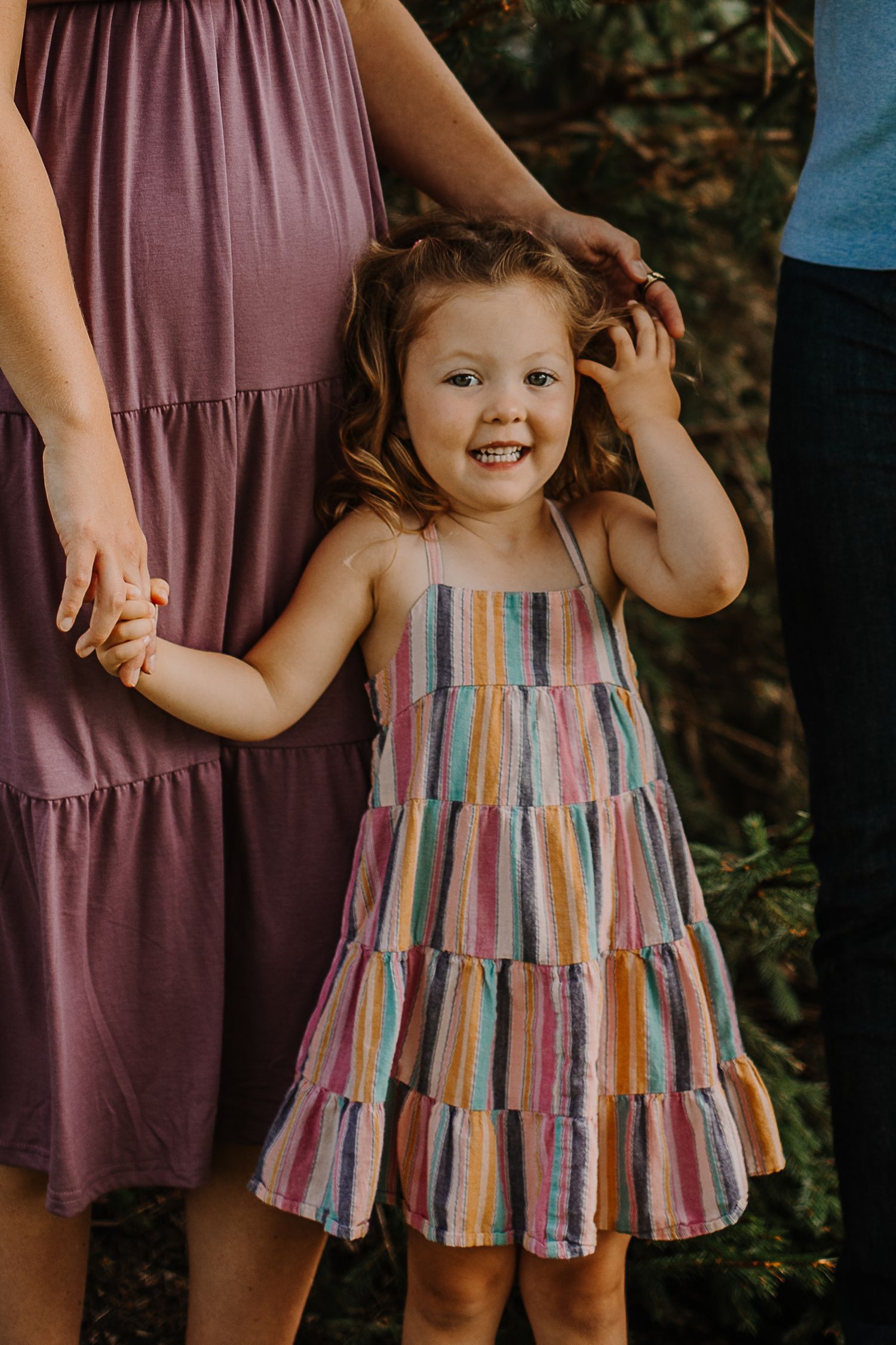 Happy little girl in colorful striped dress smiling while holding parents hands outdoors.