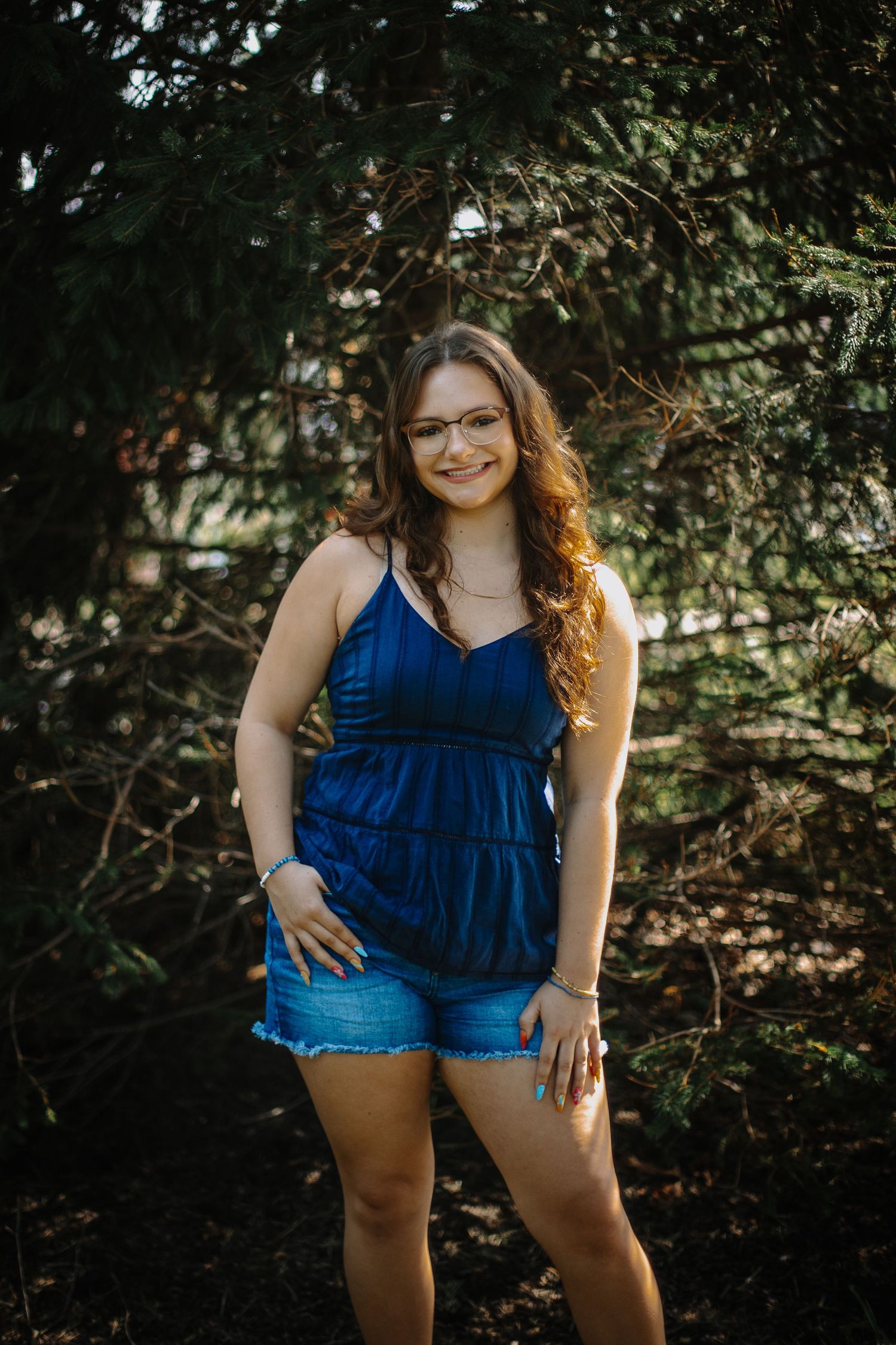Young woman in blue tank top and denim shorts smiling among evergreen trees outdoors.