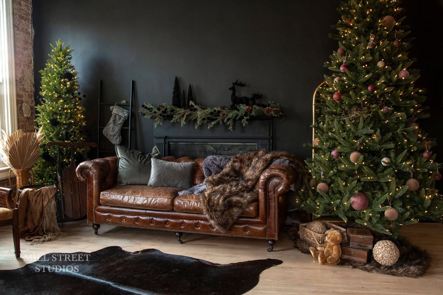 Elegant Christmas studio with leather sofa, decorated fireplace mantel, and two ornament-filled trees.