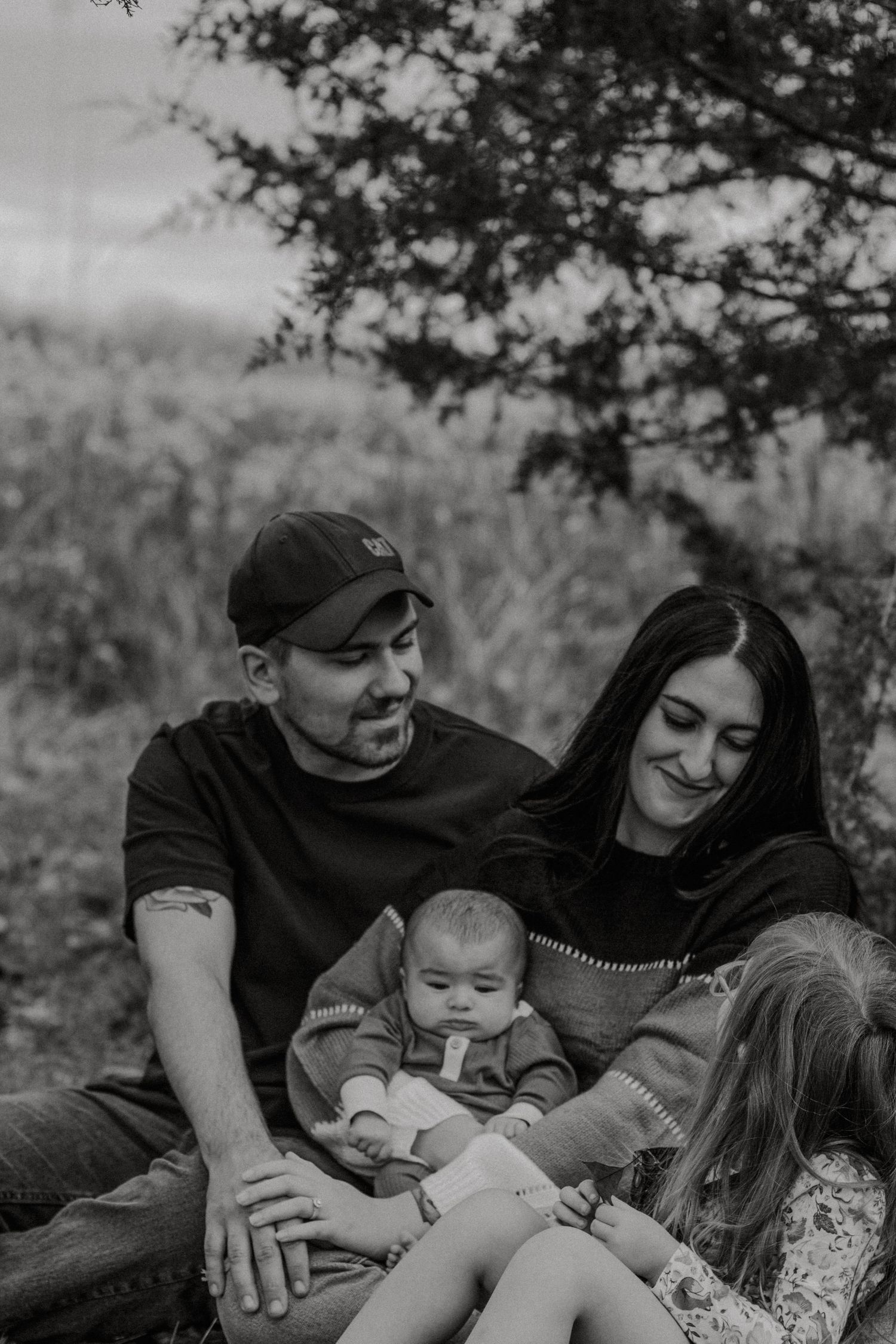 Black and white photo of a couple sitting outdoors with a baby and young child under a tree.