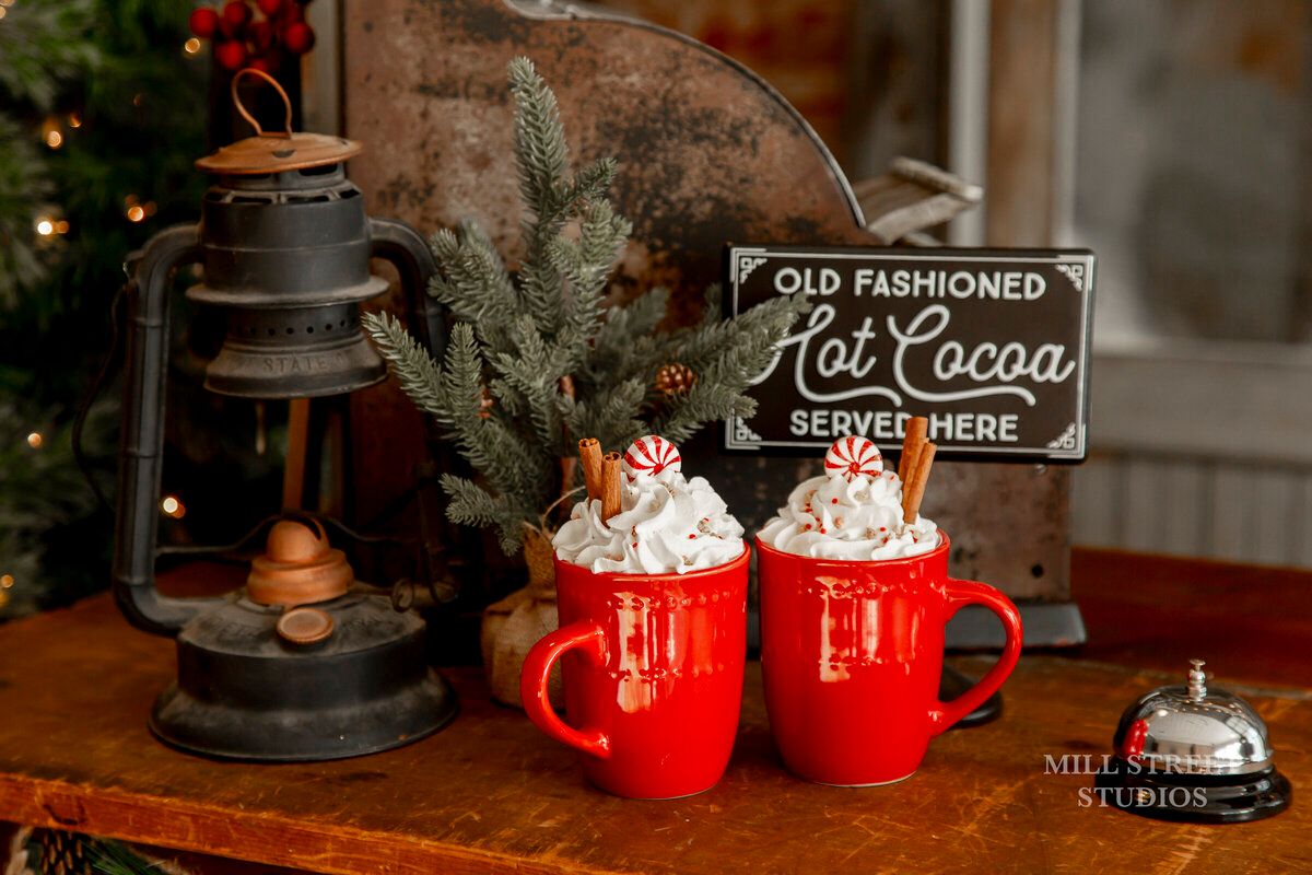 Two red mugs of hot cocoa topped with whipped cream and candy canes on a festive holiday table with vintage lantern.