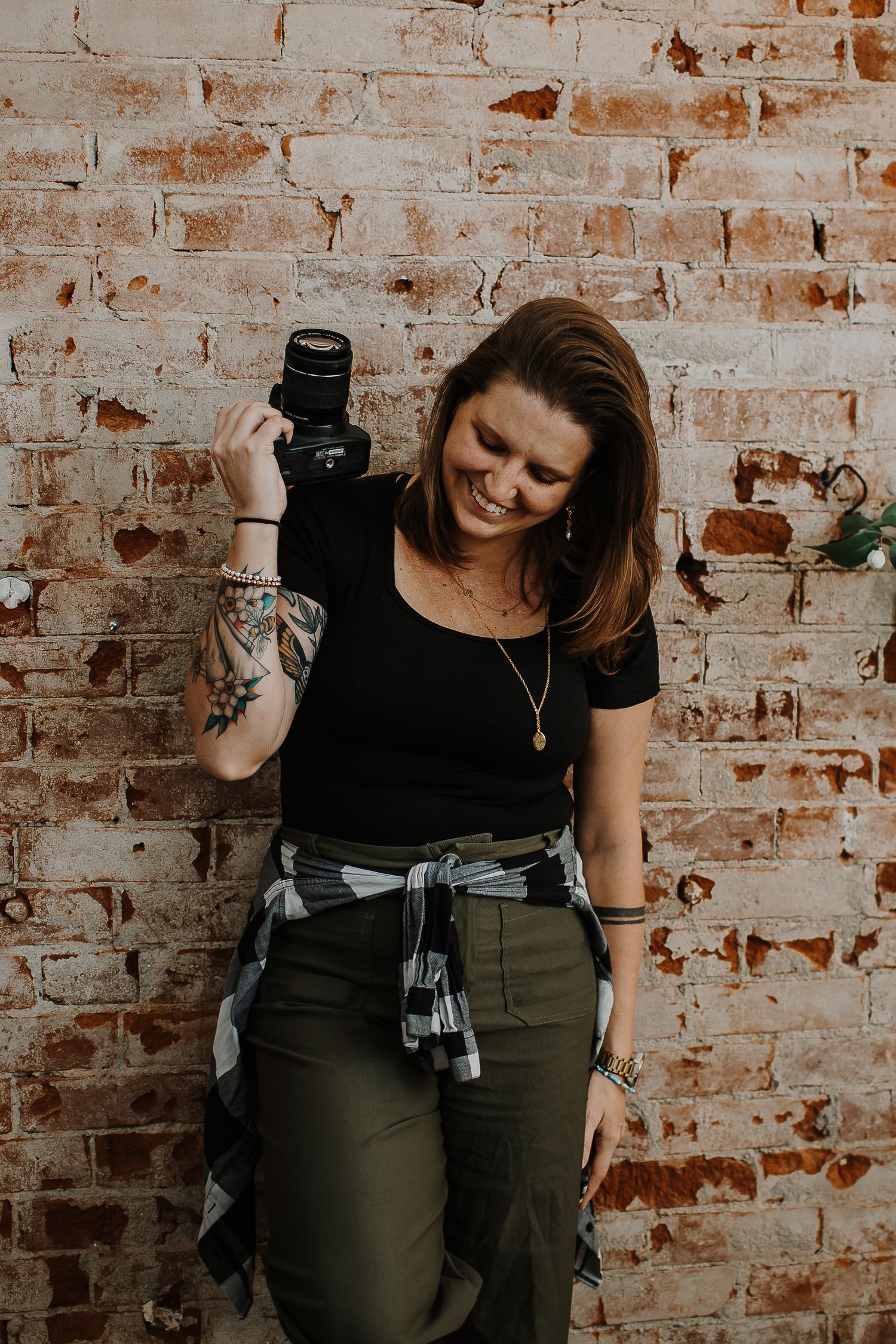 Smiling woman with tattooed arm holds a black camera against a weathered white brick wall, wearing a black top and olive pants.