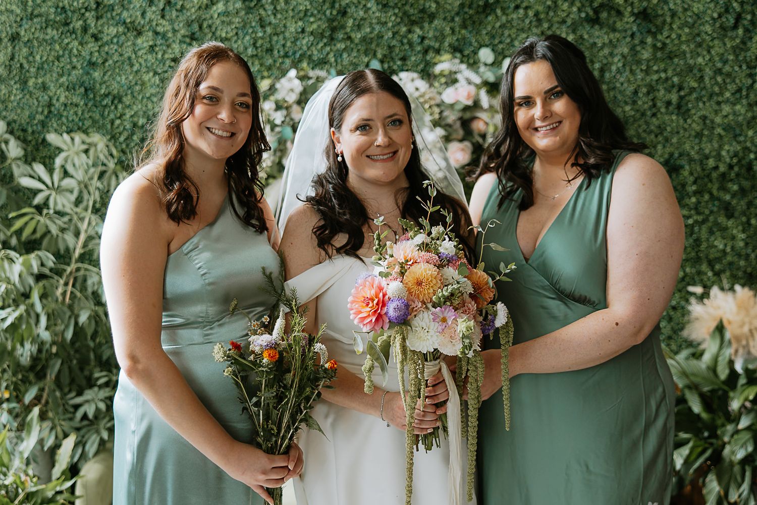 Bride in white gown holds colorful bouquet flanked by two bridesmaids in sage green dresses before lush greenery backdrop.