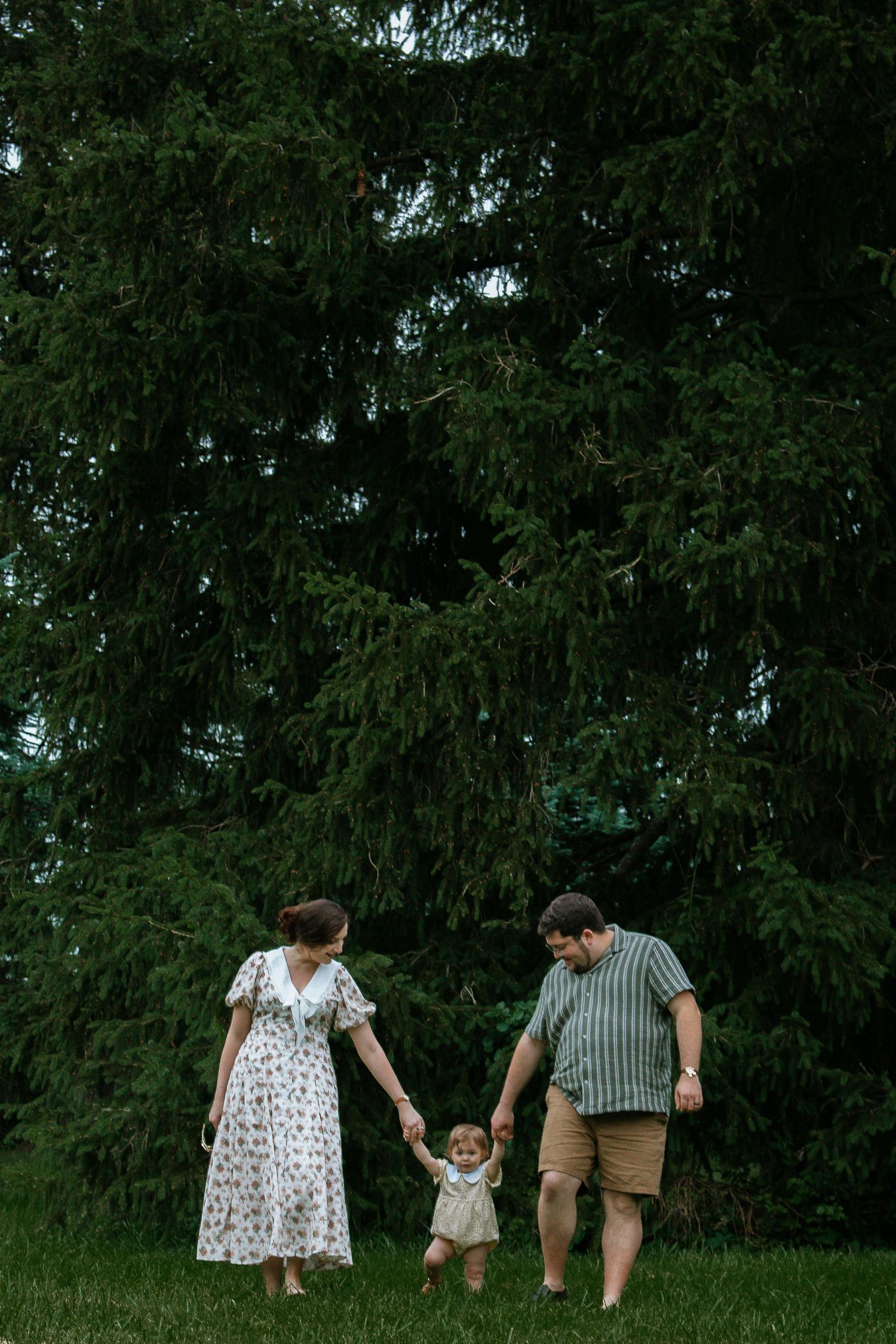 Family of three walking hand in hand on green grass with tall evergreen trees in background.