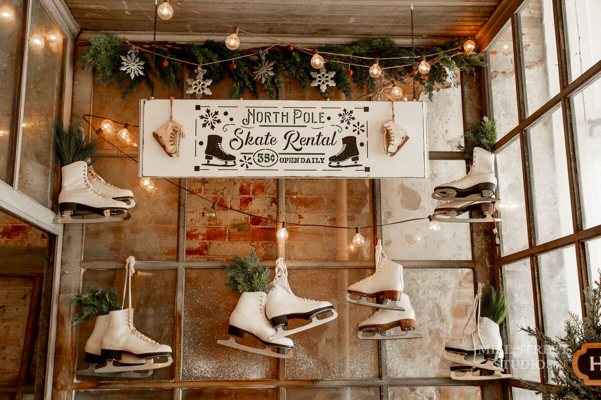 Vintage white ice skates hang on a rustic window display with a North Pole Skate Rental sign and festive holiday greenery.
