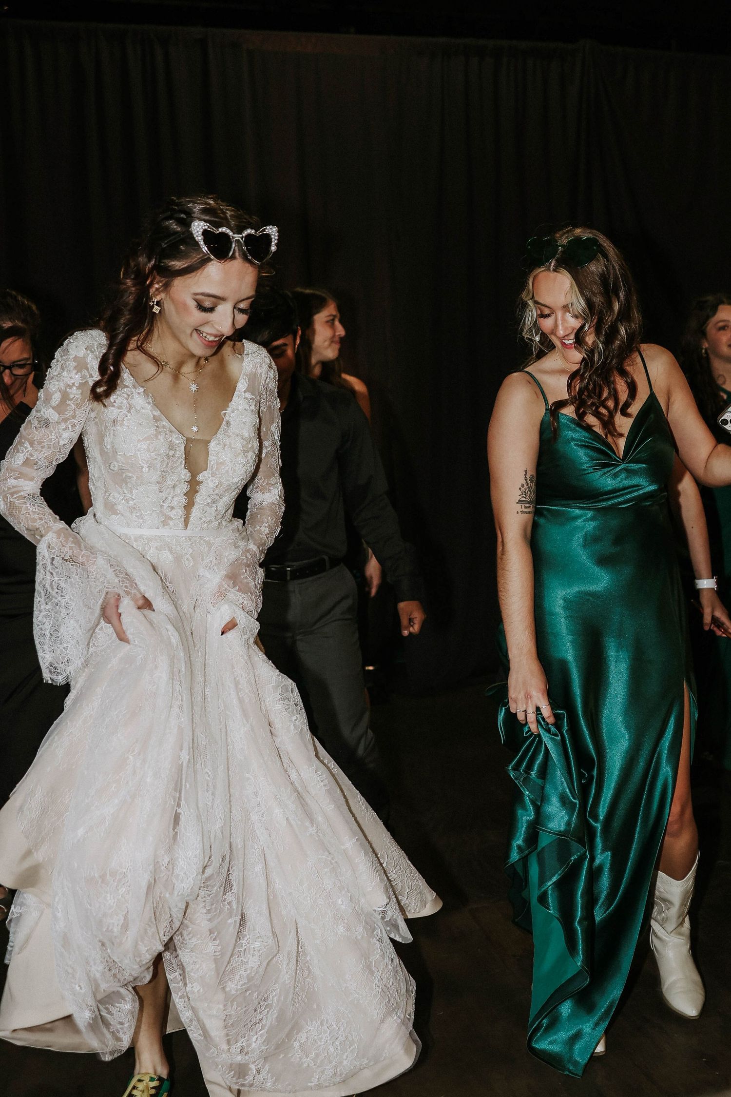 Bride in lace gown with heart sunglasses dances alongside bridesmaid in emerald satin dress at wedding reception.