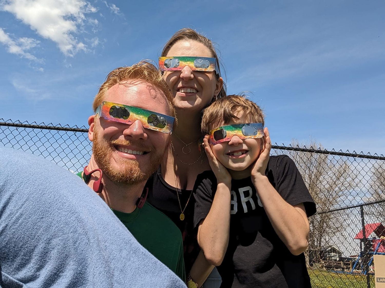 Happy family taking selfie wearing solar eclipse glasses outdoors on sunny day.