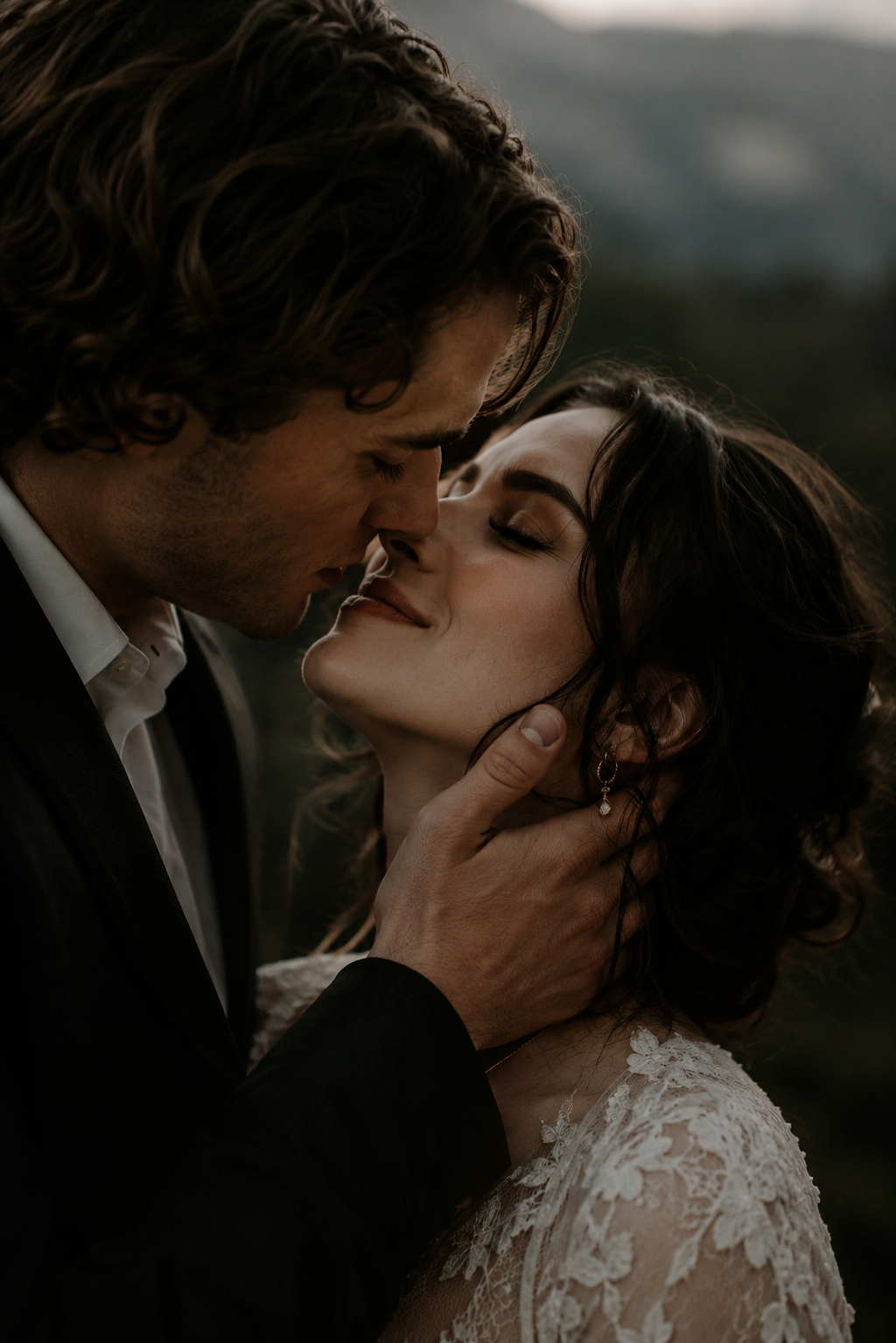 Dramatic close-up of groom gently holding bride's face for a near-kiss, bride in lace gown with mountain backdrop.