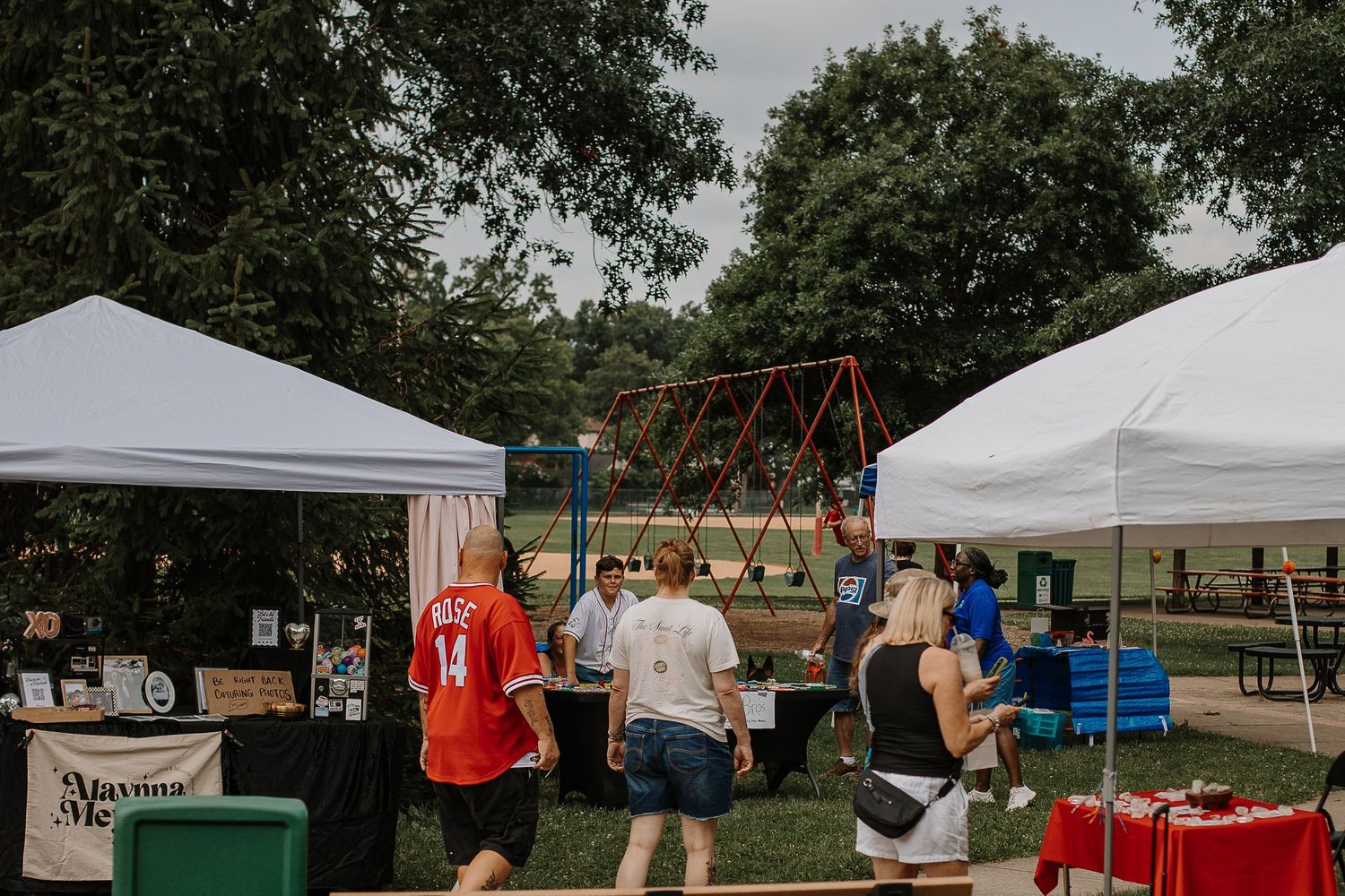 Visitors browsing vendor booths under white canopy tents at an outdoor community market with trees in the background.
