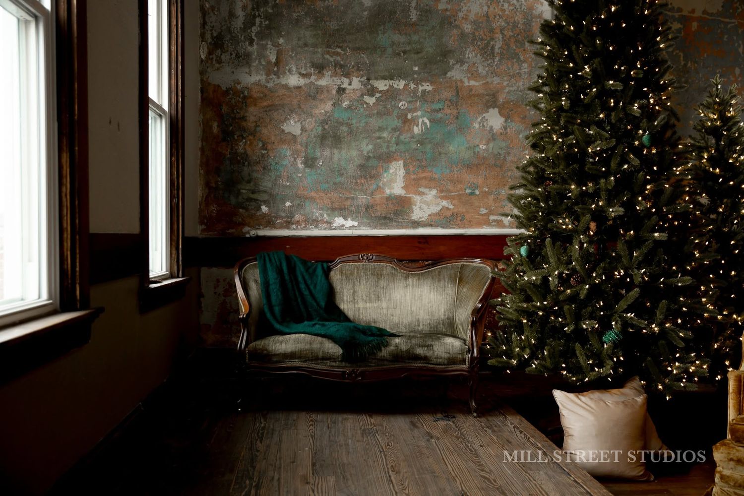 Cozy Christmas scene with a green lit tree beside a velvet sofa against a rustic distressed wall.