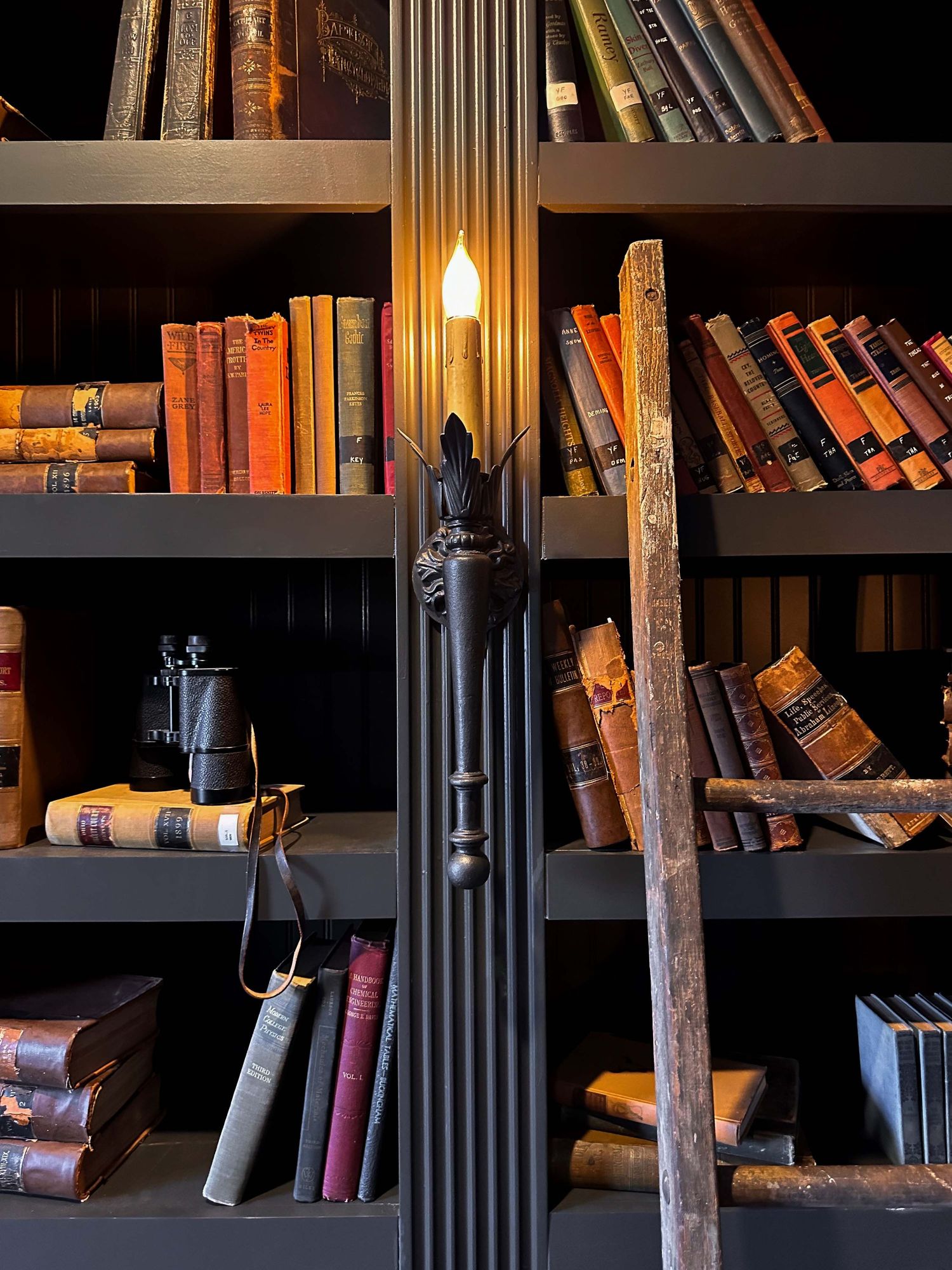 Dark wooden bookshelf filled with antique books, a glowing candle lamp, binoculars, and a rustic wooden ladder.