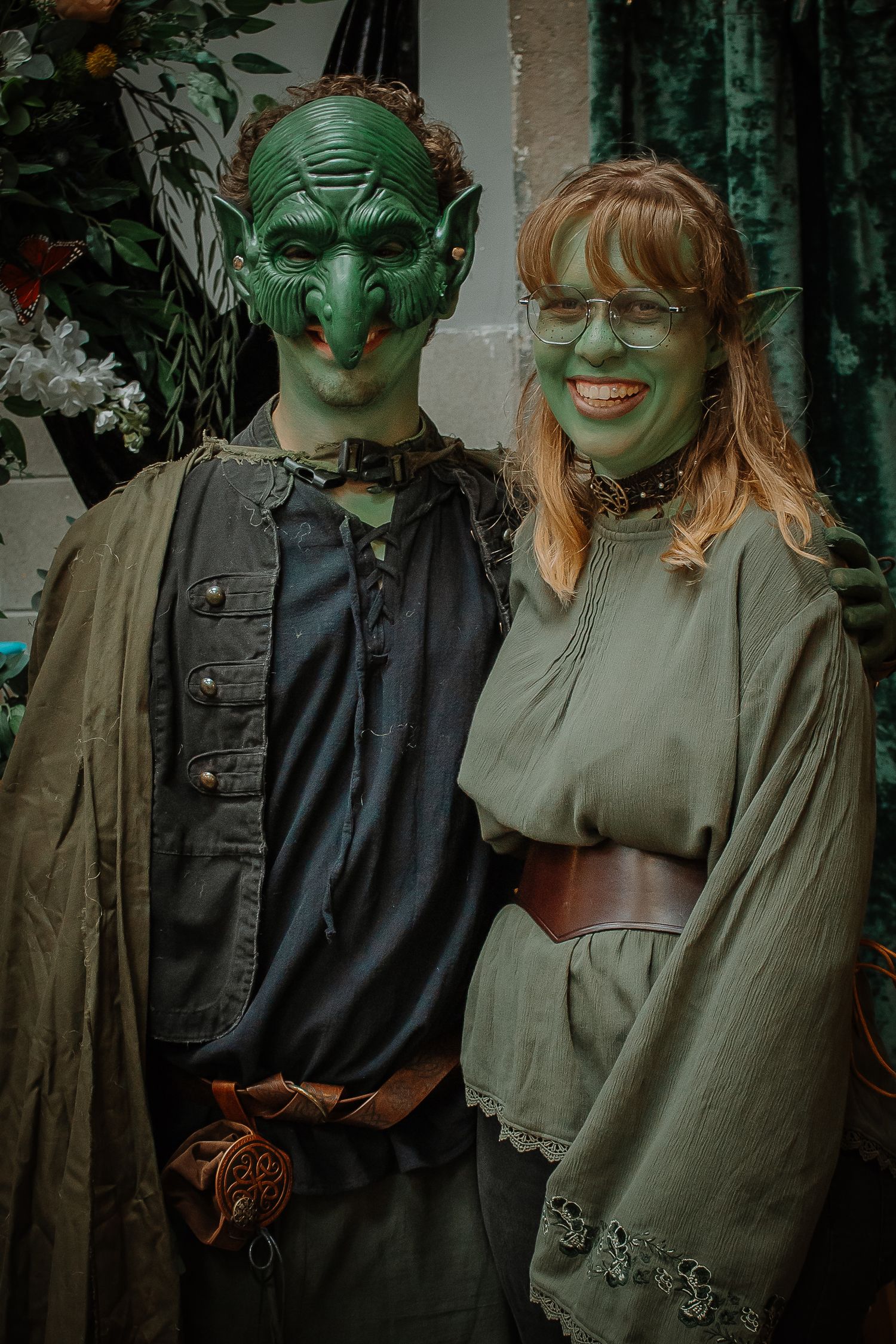 Couple posing in detailed green goblin costumes with face paint and pointed ears at a fantasy event.