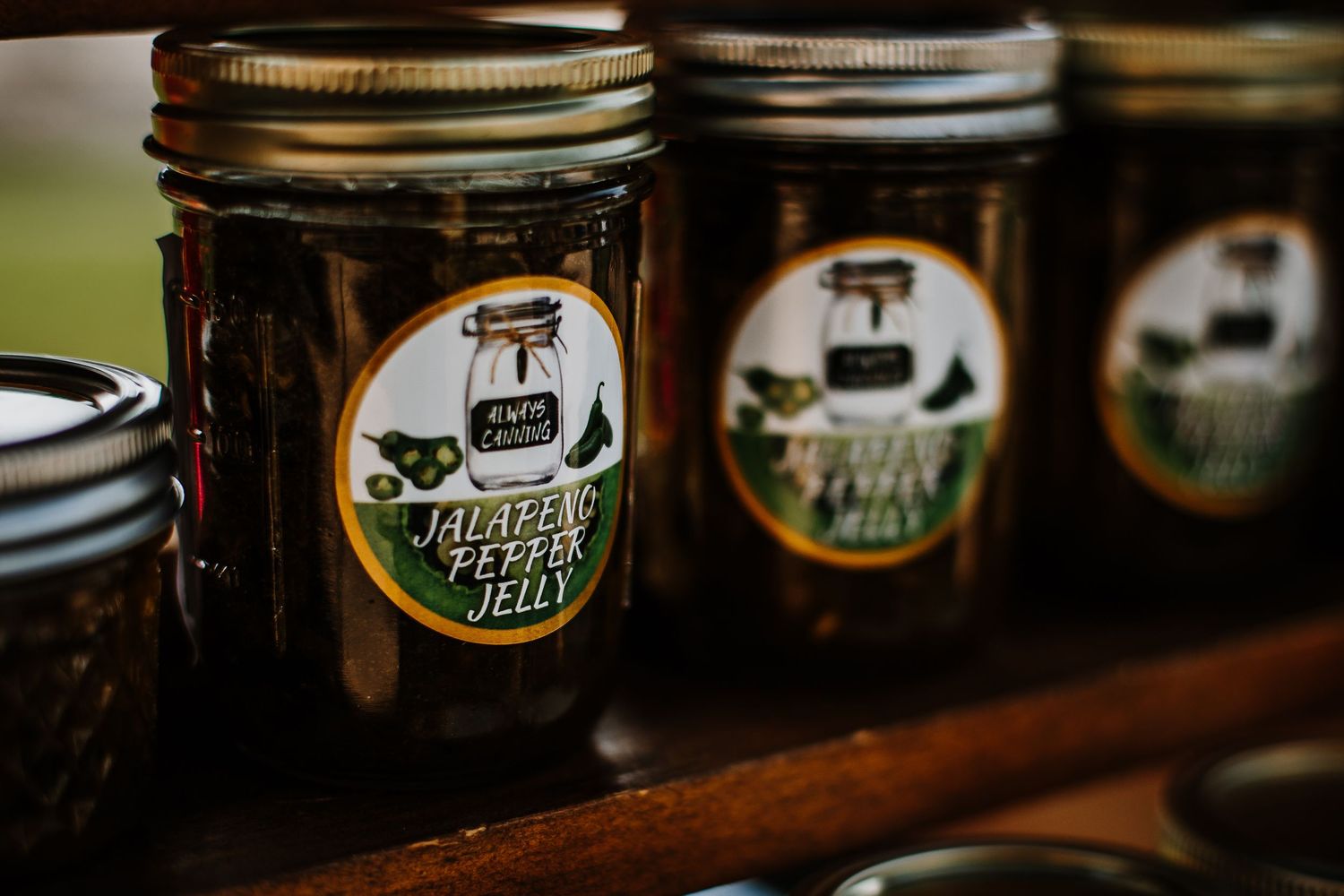 Jars of homemade jalapeno pepper jelly and other preserves on a rustic wooden shelf.