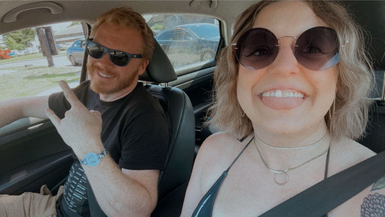 Smiling couple wearing sunglasses taking a selfie together inside a car on a sunny day.