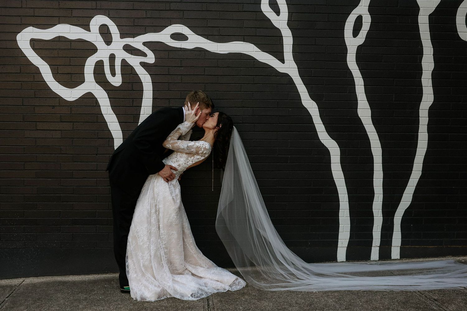 Bride and groom share a romantic kiss in front of a black wall with white floral mural, bride wearing lace gown with veil.