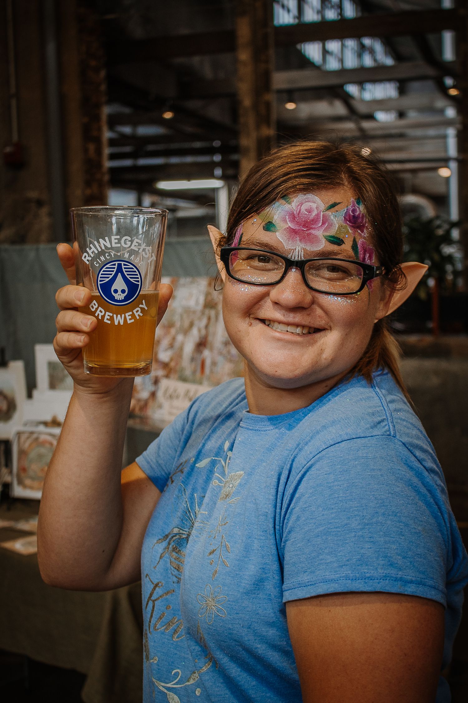 Smiling woman with elf ears and floral face paint holding a Rhinegiest Brewery beer glass at an event.