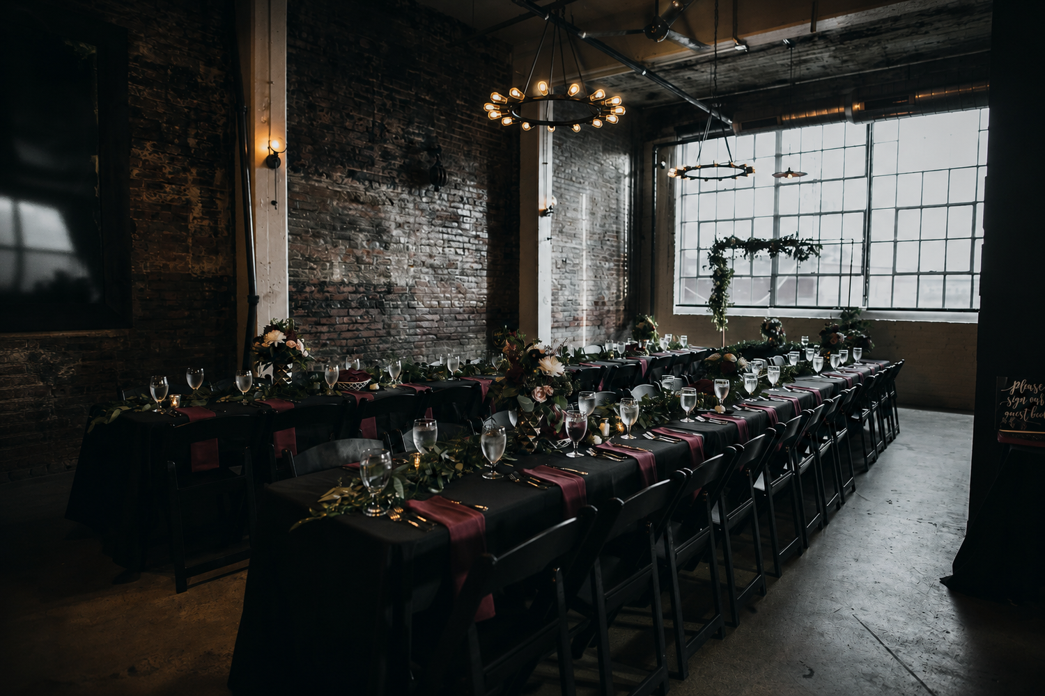 Elegant industrial wedding reception with long black tables, string lights, and brick walls in a moody venue.