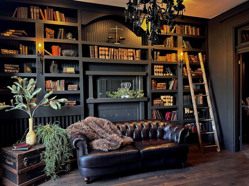 Dark moody home library with leather Chesterfield sofa, floor-to-ceiling bookshelves, wooden ladder, and chandelier.