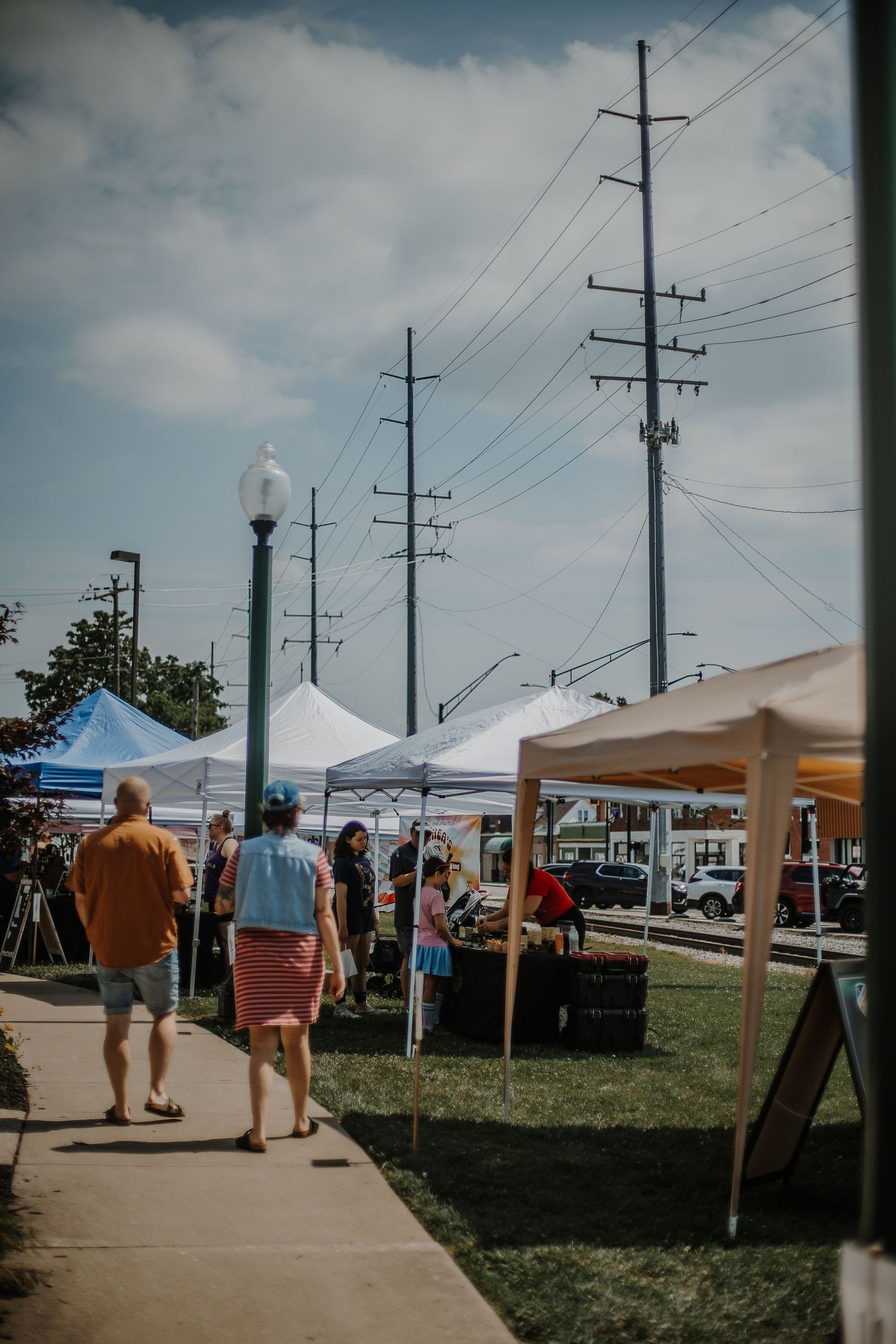People walking along a sidewalk at an outdoor farmers market with white canopy tents and utility poles under cloudy skies.