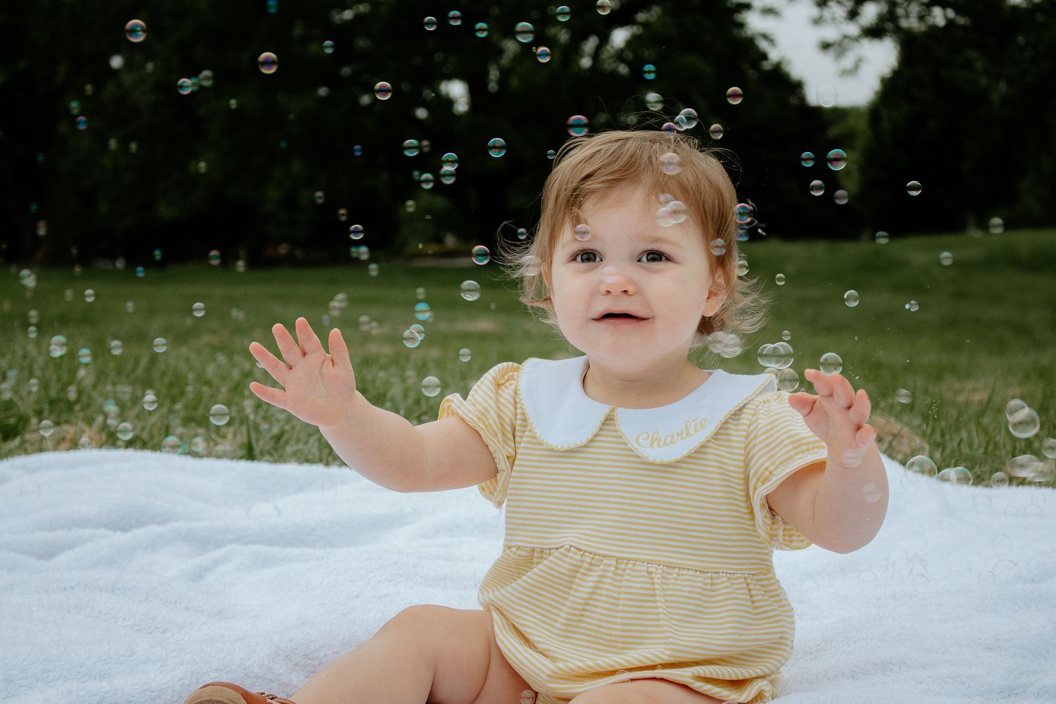 A delighted baby girl in a yellow romper reaches for floating bubbles while sitting on a white blanket outdoors.