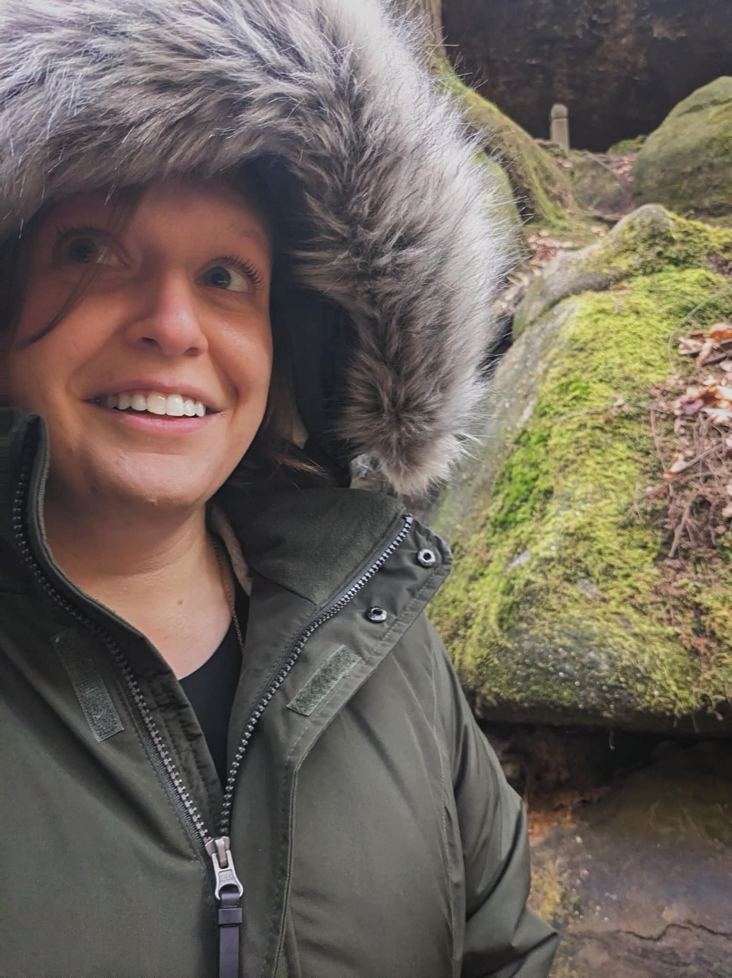 Woman smiling in fur-hooded green parka near mossy rocks outdoors.