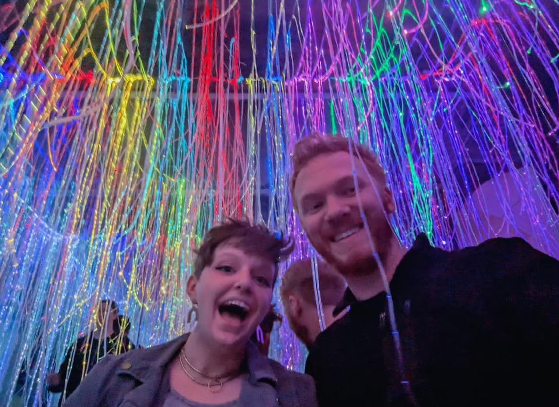 Happy couple smiling under colorful rainbow LED light installation at an art exhibit.