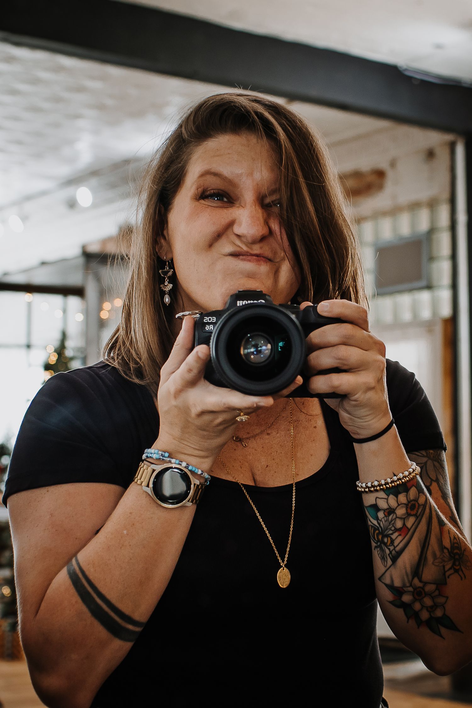 Woman with floral arm tattoos takes a mirror selfie holding a mirrorless camera while squinting playfully in a bright, rustic studio.