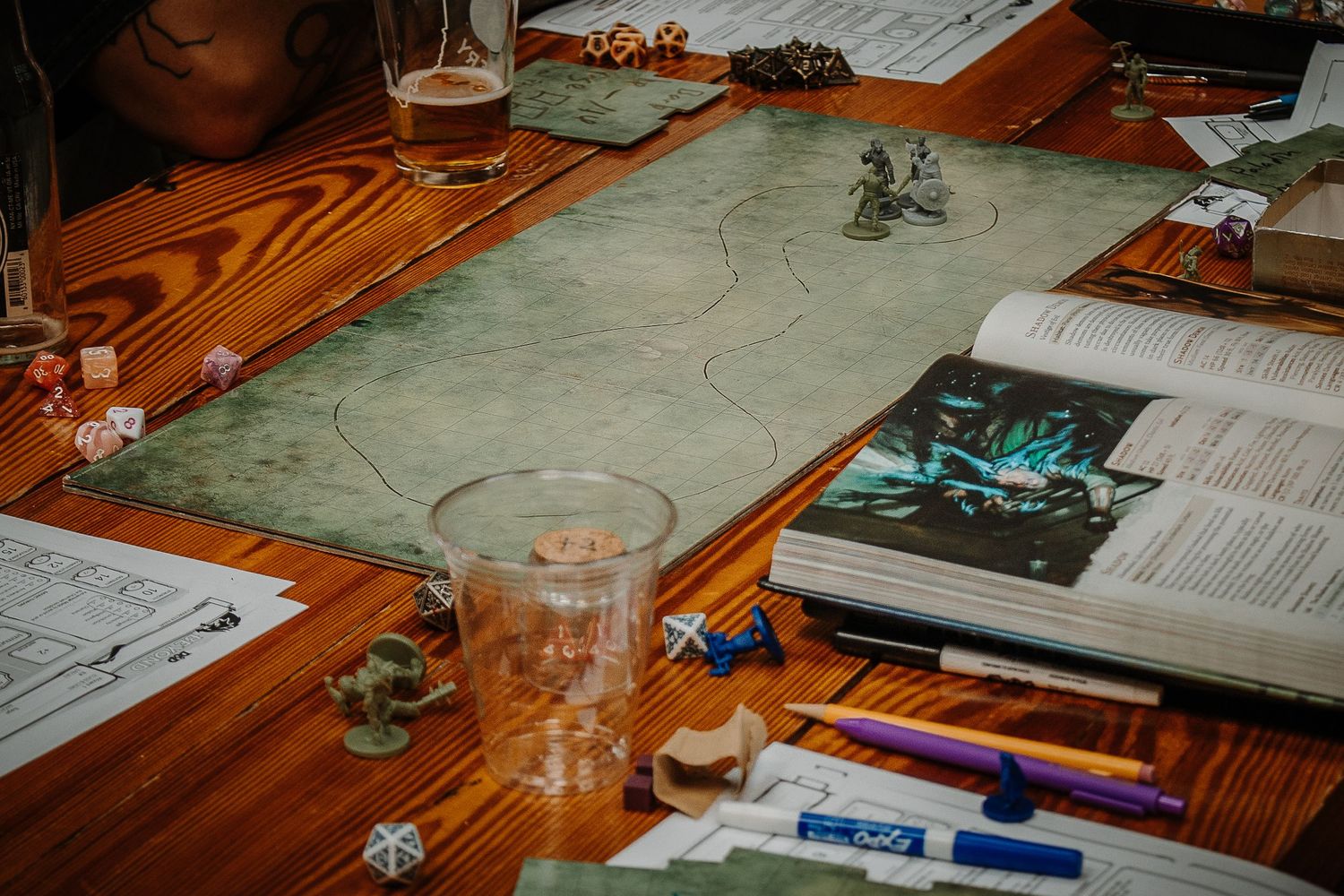A tabletop RPG setup with a battle map, dice, miniatures, rulebook, and drinks on a wooden table.
