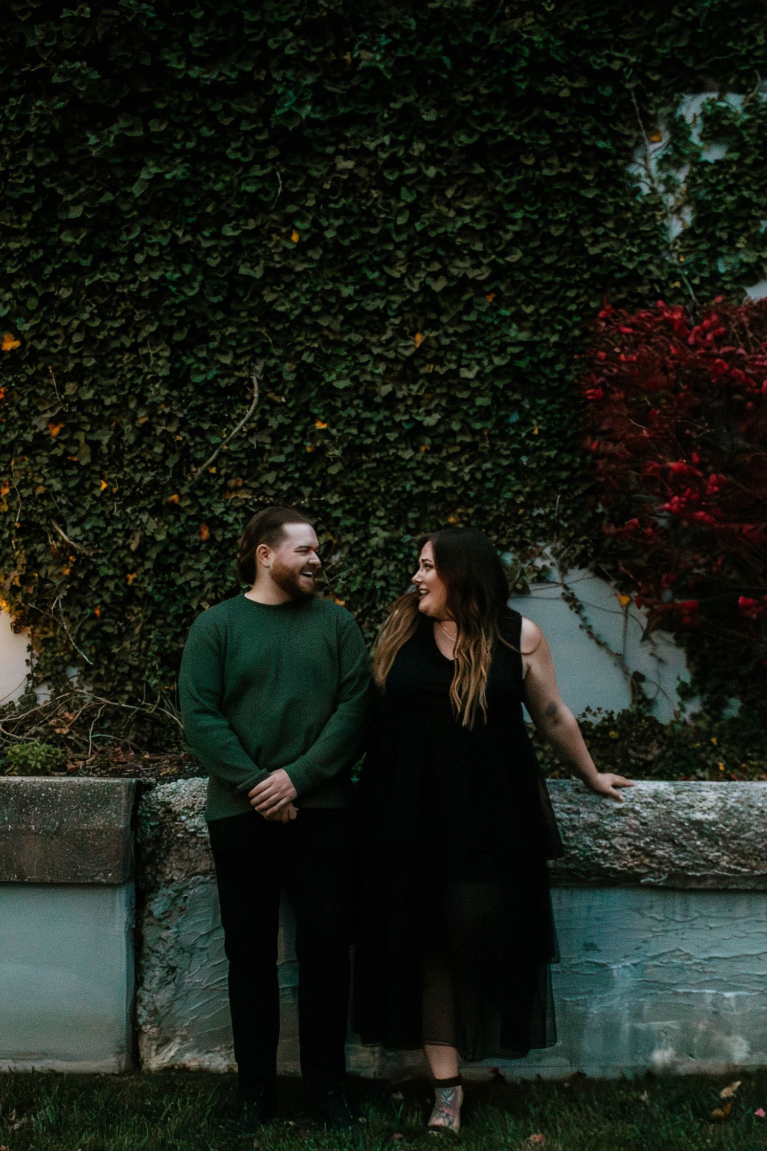 Smiling couple holding hands in front of lush ivy wall during fall engagement session.