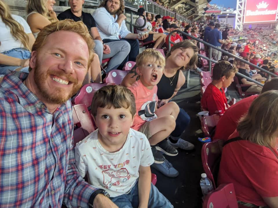 Family smiling and posing for a selfie in stadium bleachers during a live sporting event.