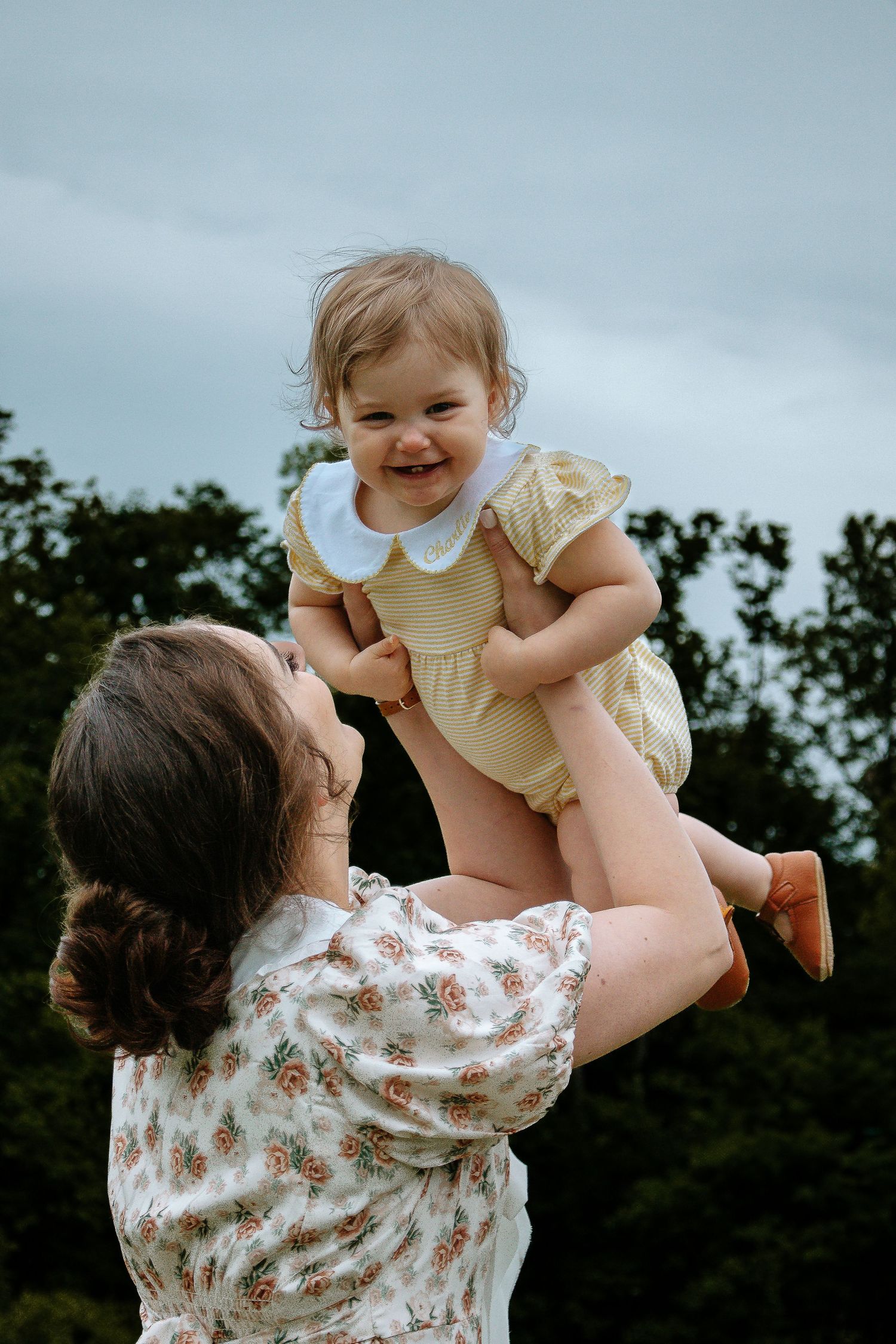 Joyful mother lifting laughing baby girl in yellow dress against a cloudy sky outdoors.