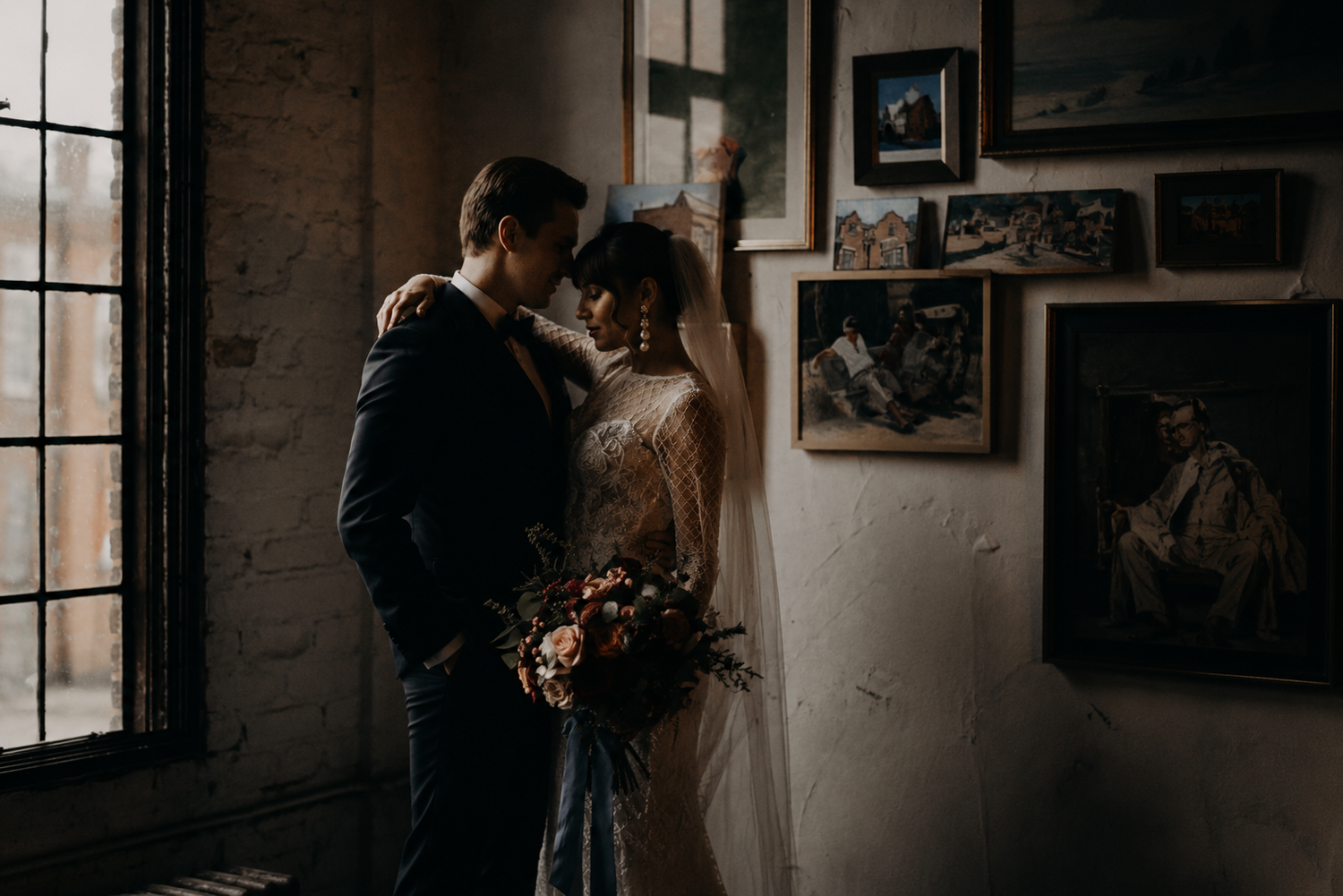 Bride and groom share intimate moment in moody industrial venue with vintage artwork and window light.