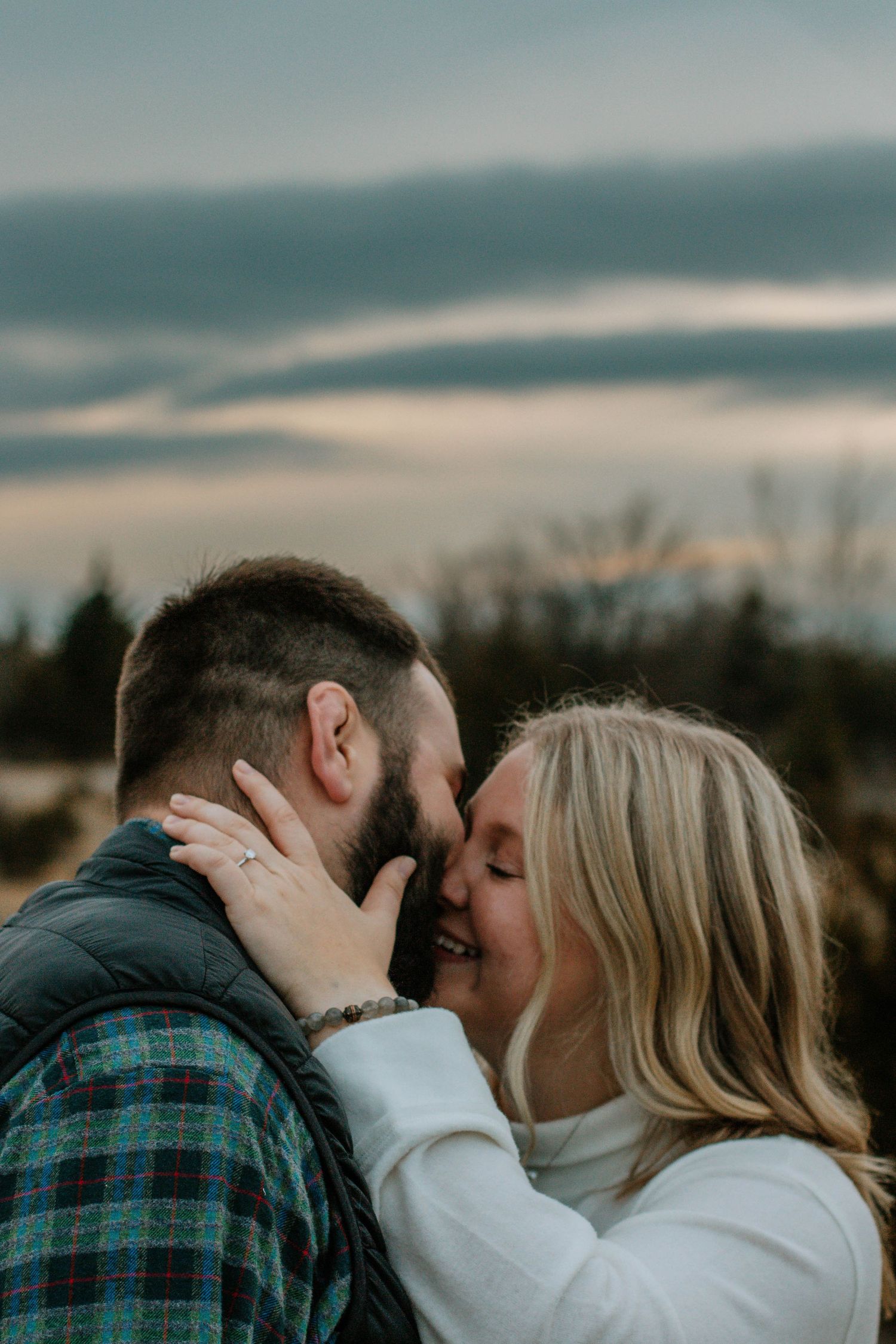 Couple sharing a romantic almost-kiss at sunset with dramatic cloudy sky backdrop during outdoor engagement session.
