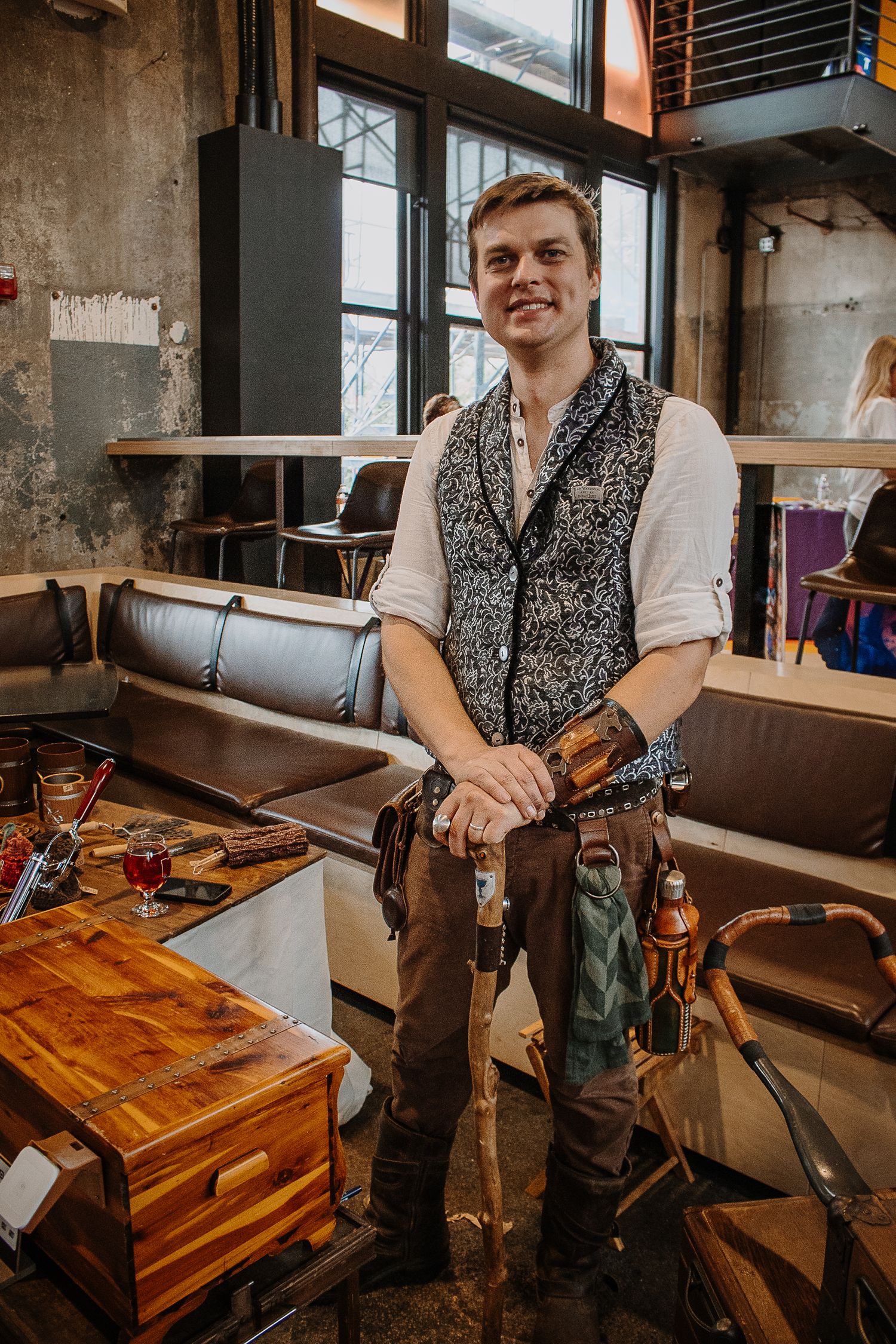 Young man in steampunk vest and tool belt poses in an industrial-style venue at a cosplay event.
